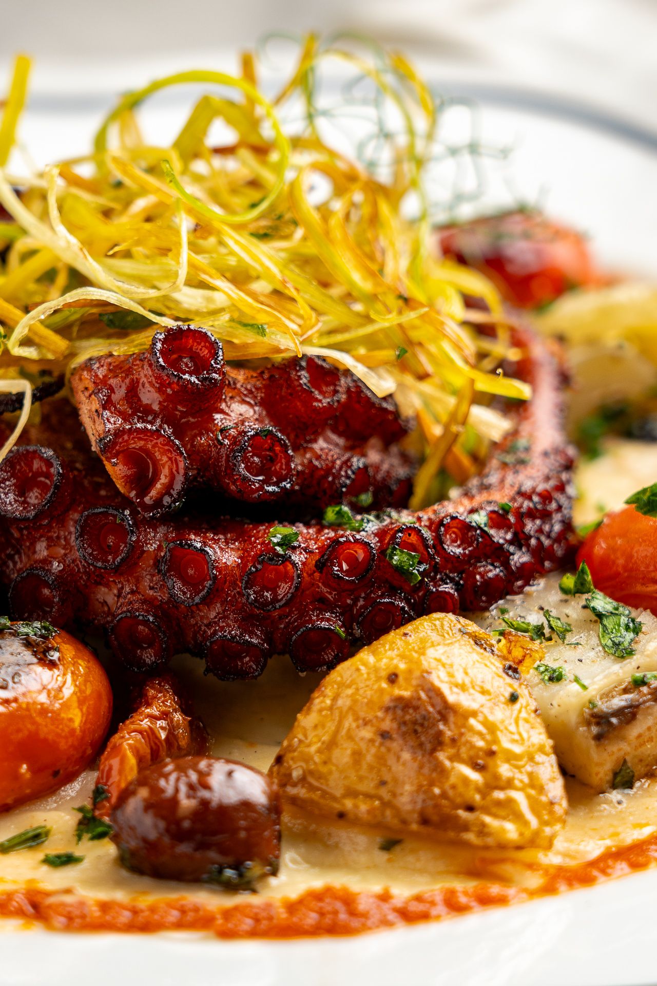 Grilled octopus tentacles served with roasted potatoes, cherry tomatoes, and crispy fried leeks on a white plate.