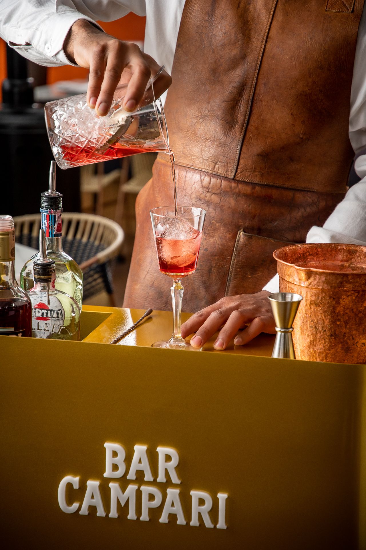 Bartender in leather apron pouring red Campari cocktail at a gold Bar Campari station with bottles nearby.
