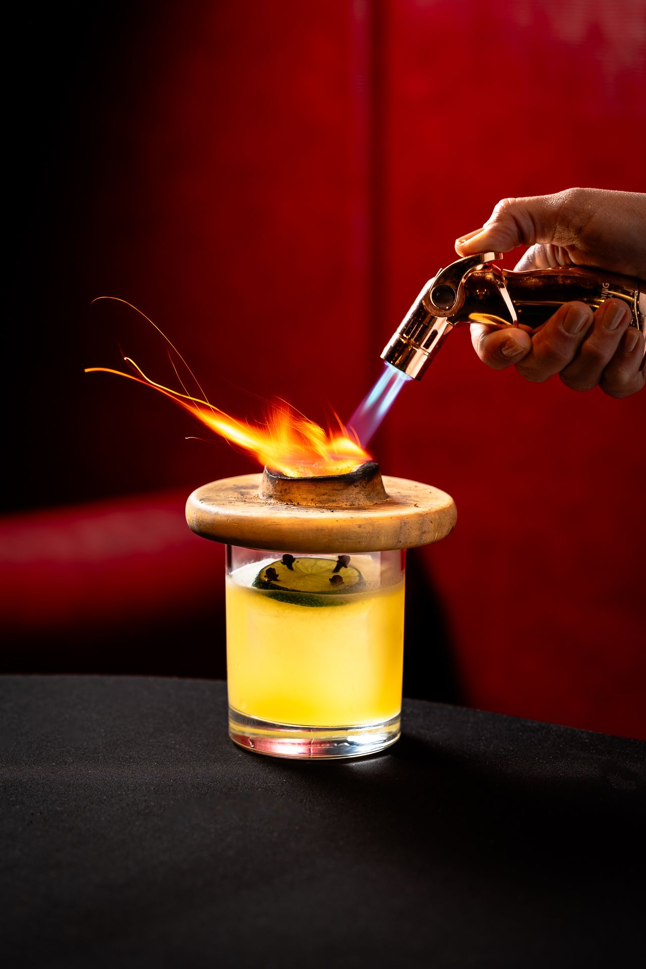 A bartender uses a blowtorch to flame a cocktail garnish on a wooden disc over a yellow drink in a glass.