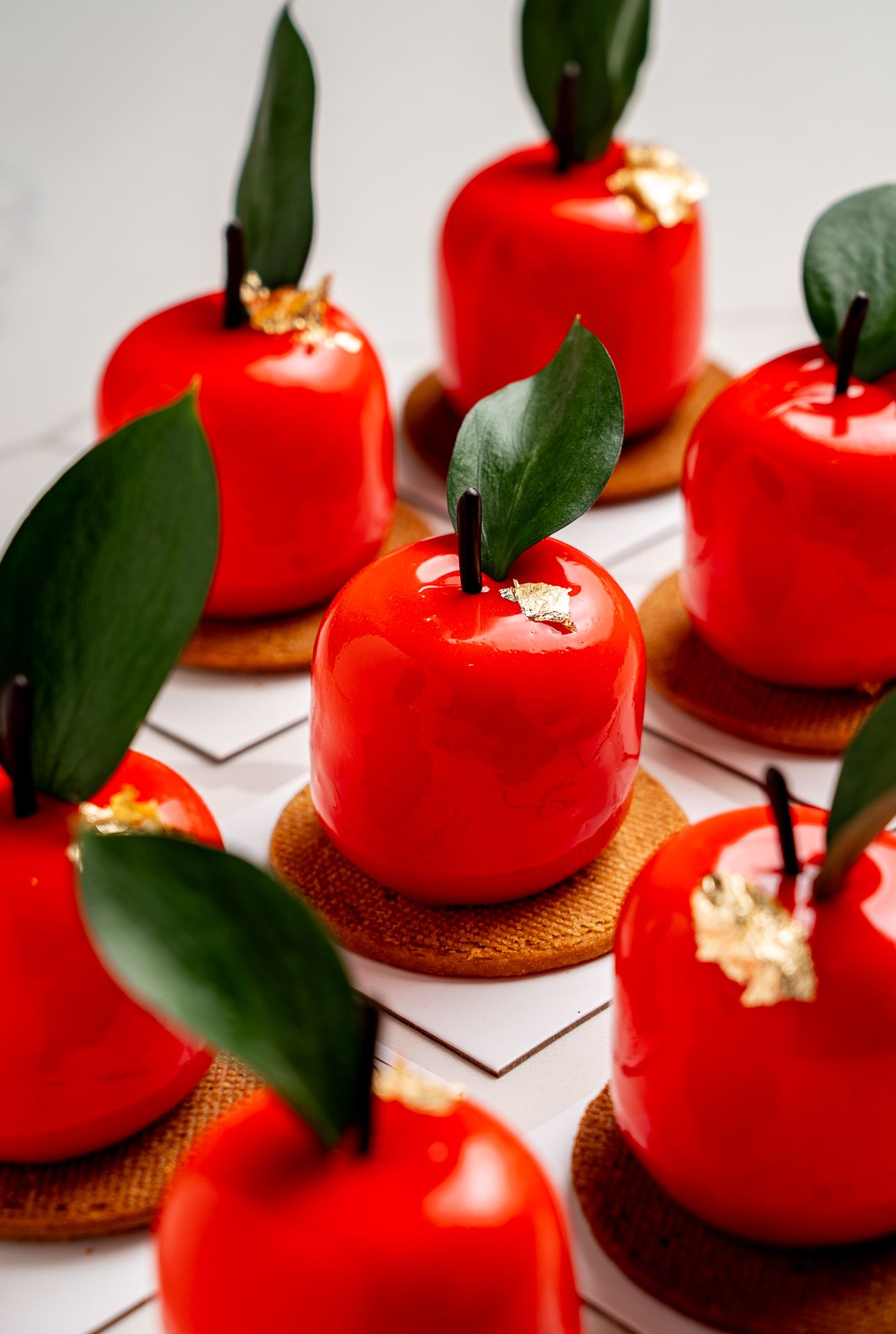 Glossy red apple-shaped mousse cakes on cookie bases, decorated with green leaves and gold flakes.