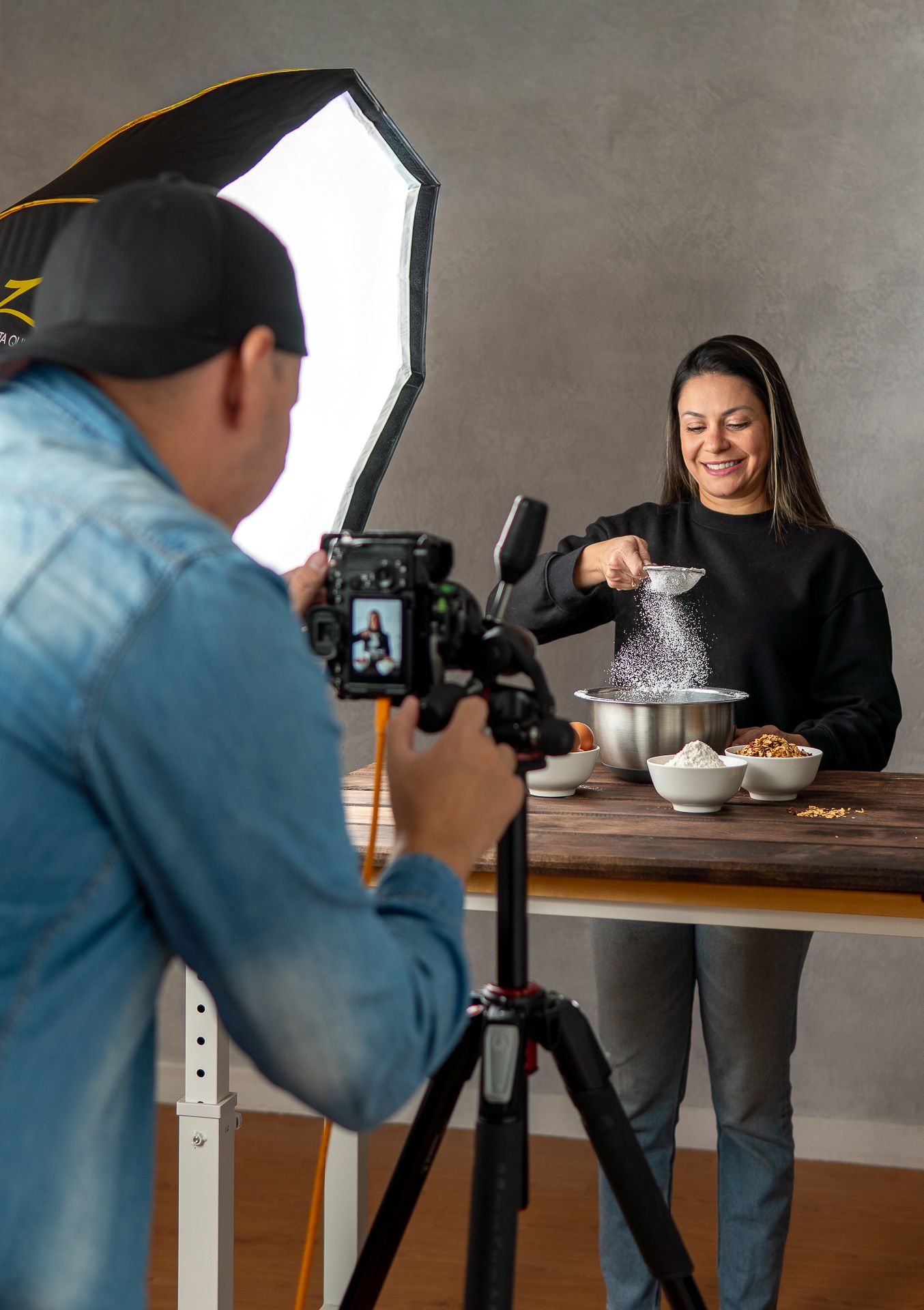 Photographer capturing a smiling woman sifting flour over bowls on a wooden table in a professional studio setup.