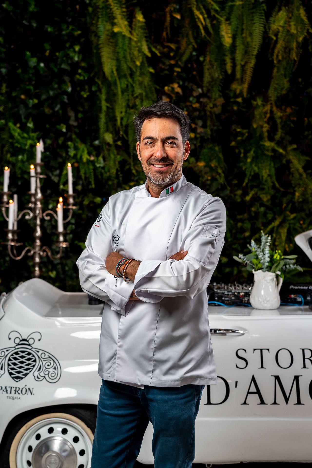 Smiling chef in white coat stands arms crossed near a vintage car with Patron branding outdoors.