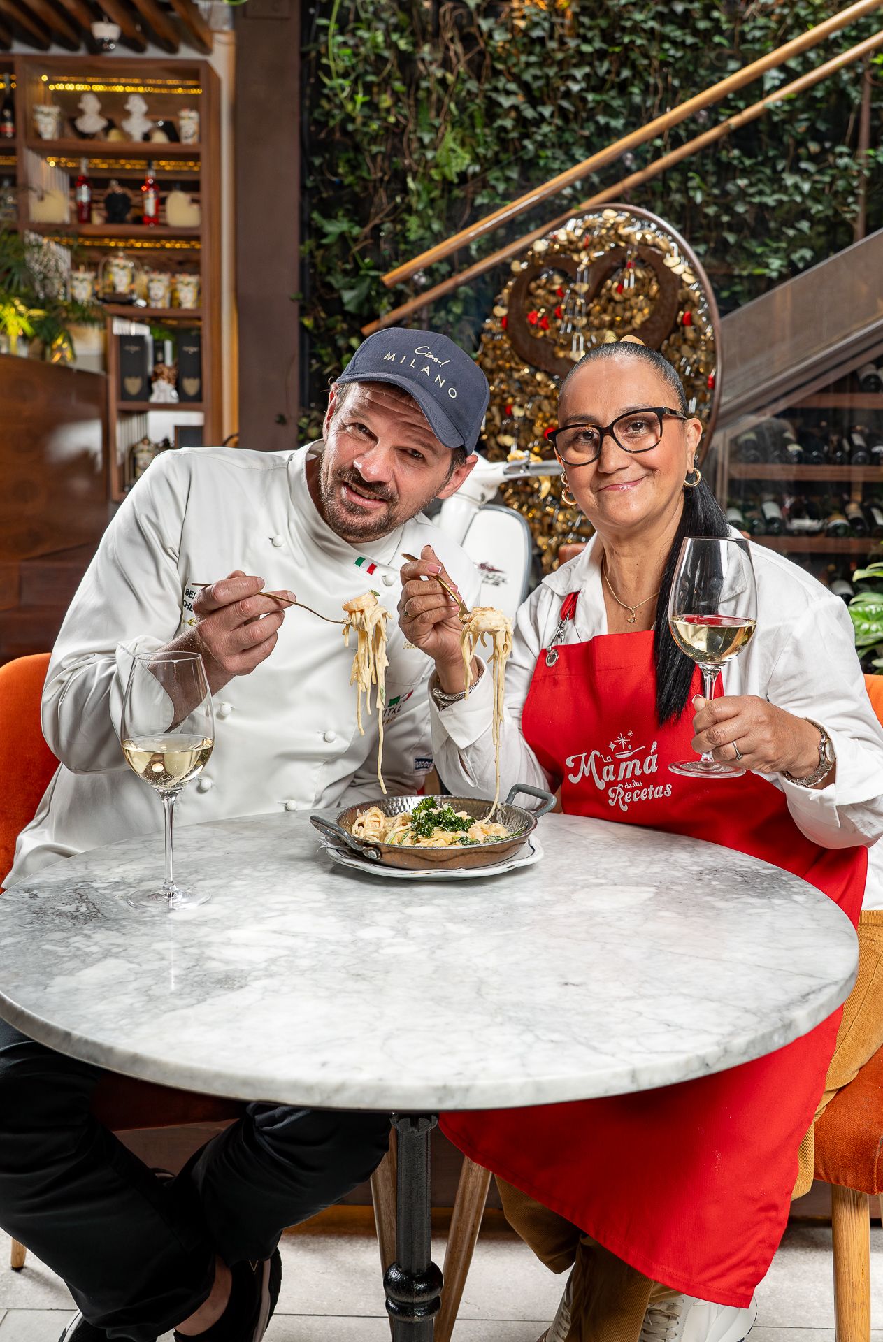 Two chefs smiling at a restaurant table, enjoying pasta and white wine, wearing chef whites and a red apron.