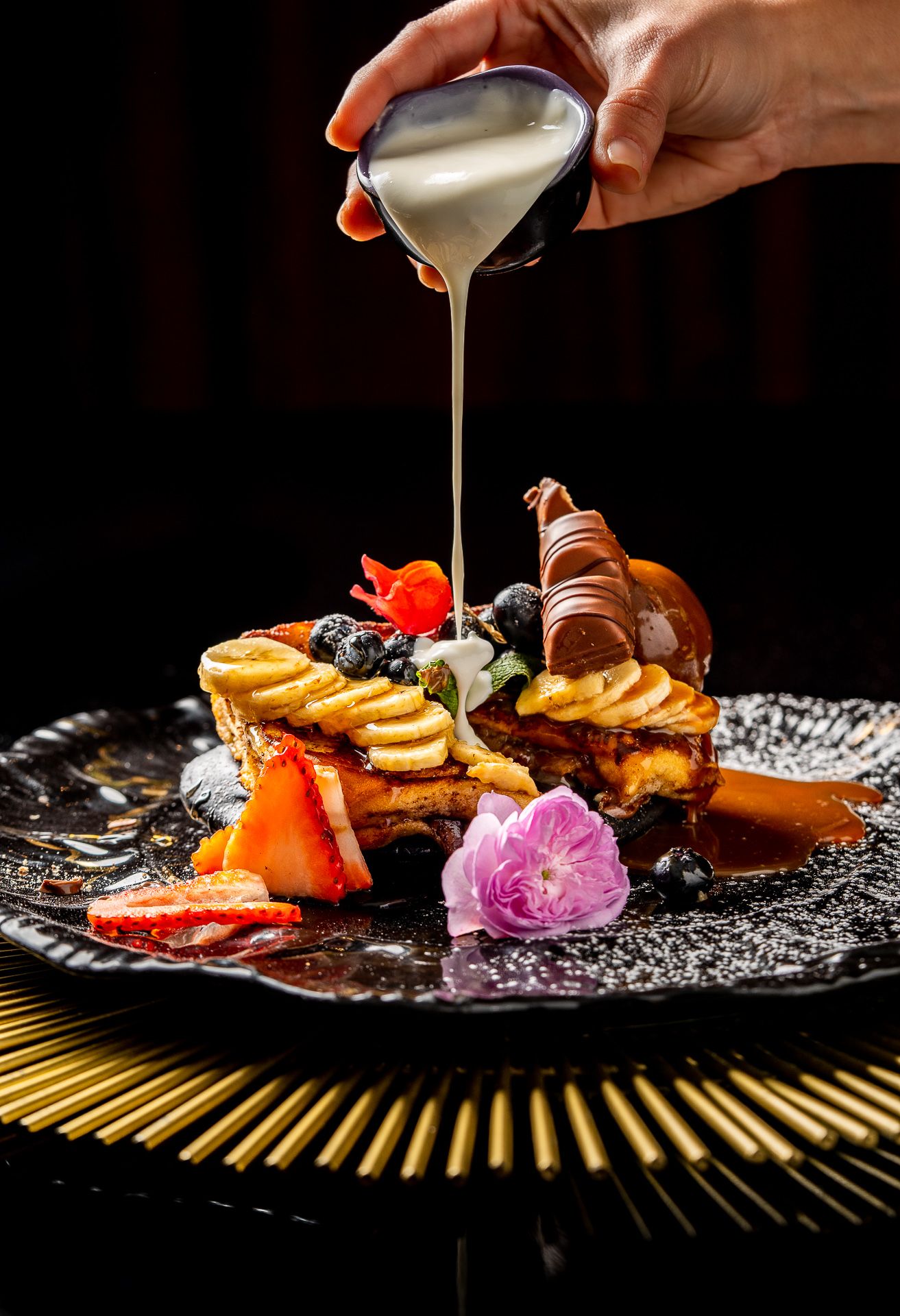 Cream being poured over an elegant dessert plate with fresh berries, strawberries, banana, and a pink flower.