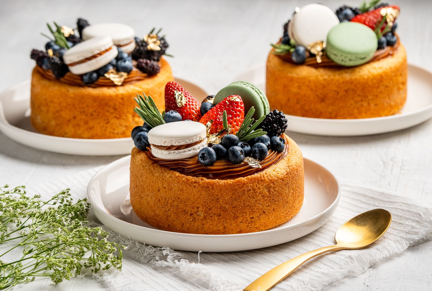 Three elegant chiffon cakes topped with fresh strawberries, blueberries, macarons, and blackberries on white plates.