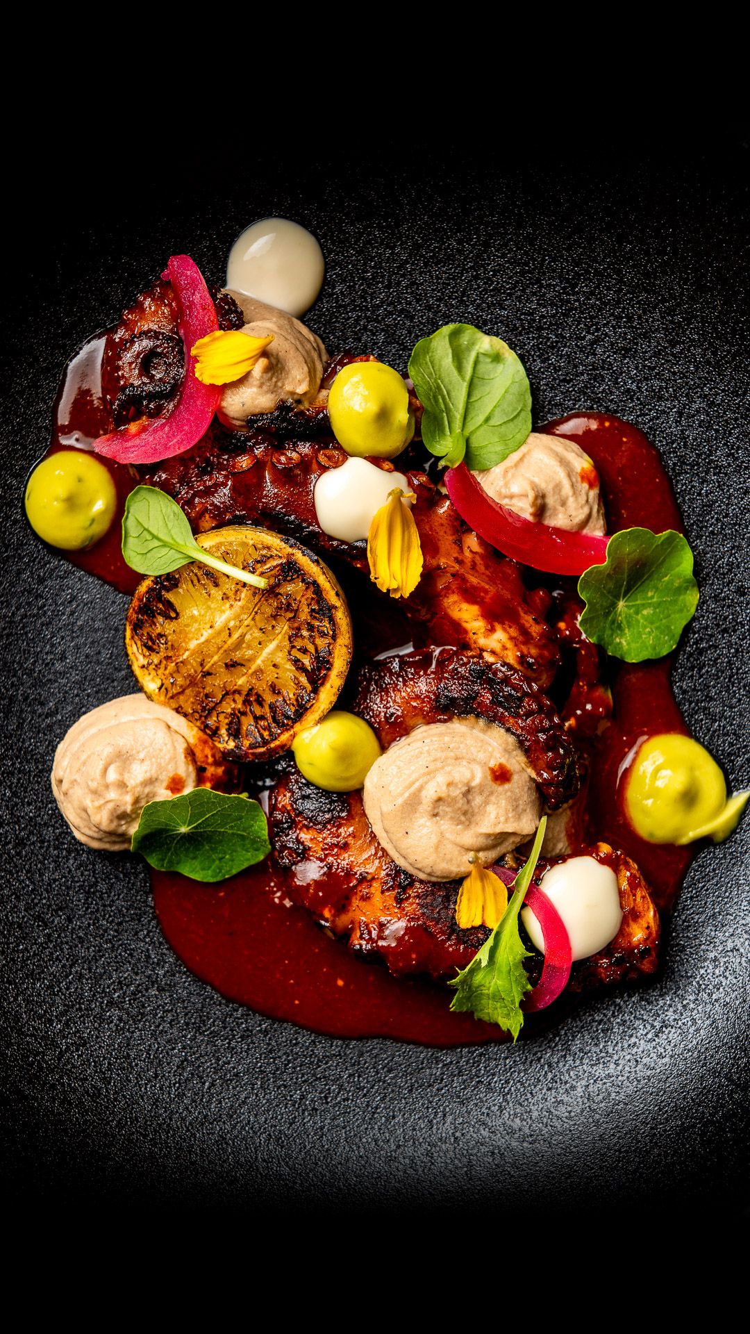 Elegant fine dining dish with glazed meat, olive spheres, cream dollops, edible flowers, and greens on a dark plate.