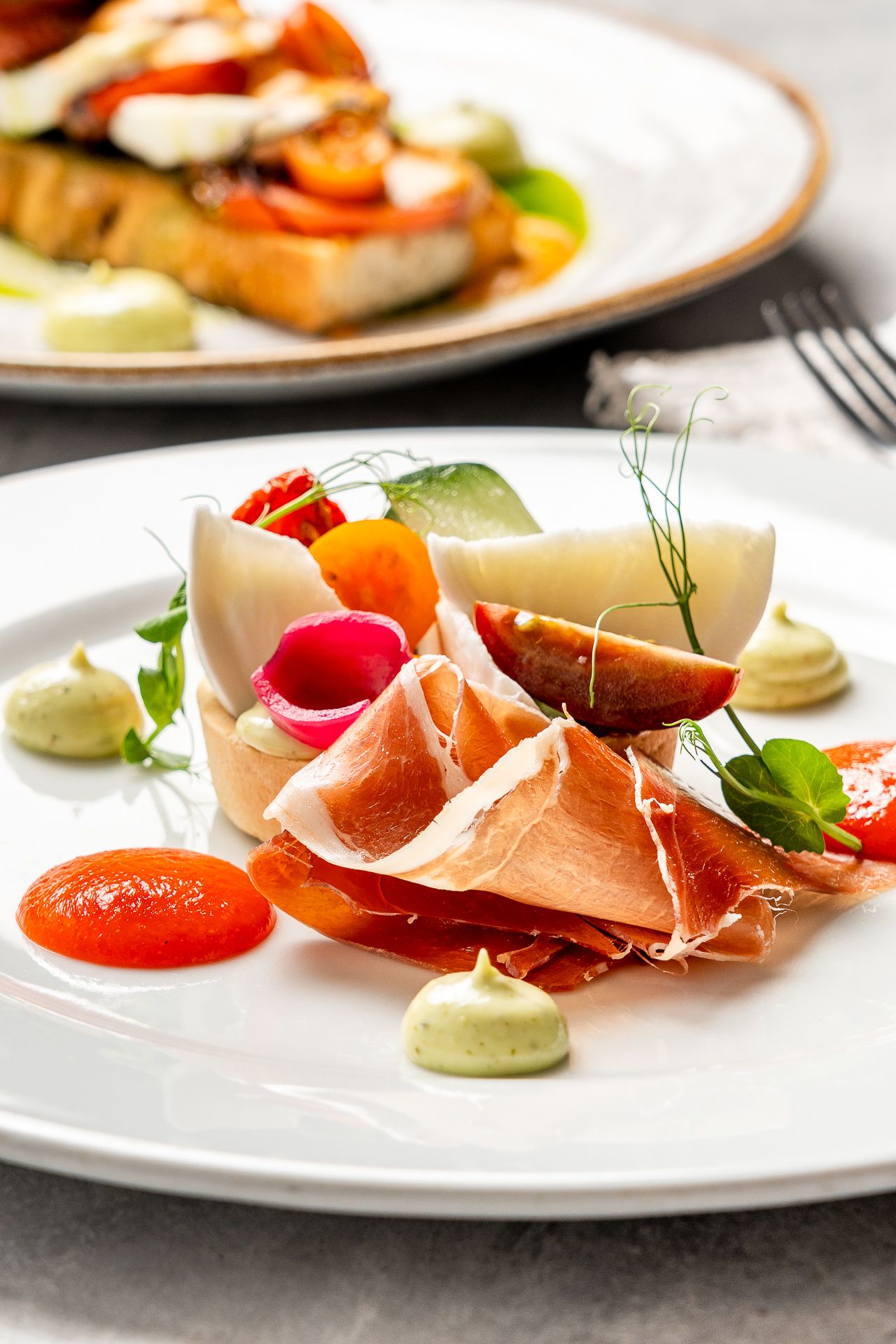 Elegant prosciutto appetizer plate with fresh vegetables, avocado mousse, and tomato sauce on white ceramic dish.