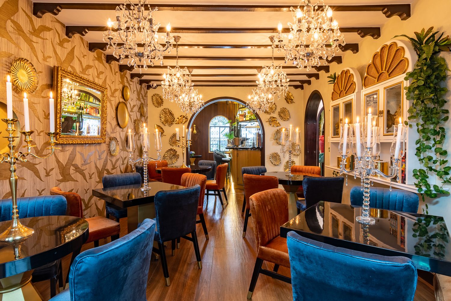 Elegant restaurant interior with crystal chandeliers, blue velvet chairs, dark wood tables, and ornate gold wallpaper decor.