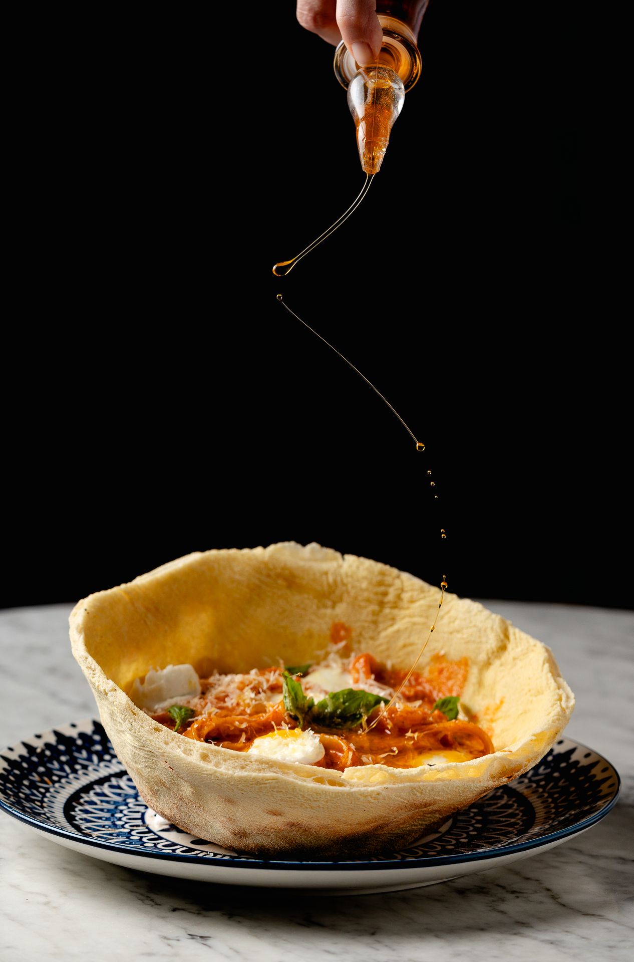 Honey being drizzled from a small bottle into a crispy tortilla bowl filled with tomato sauce, cheese, and fresh basil.
