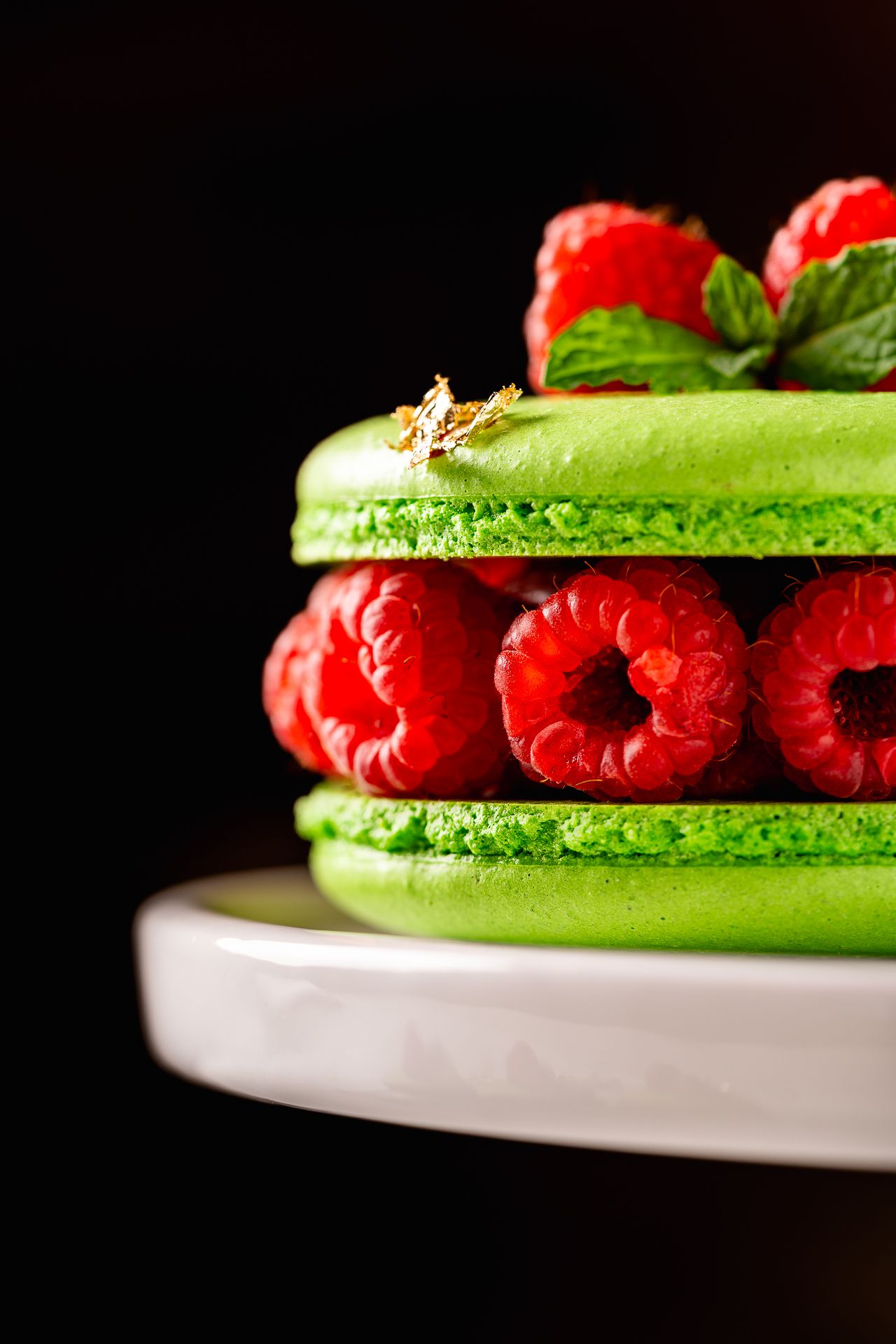 Green macaron cake with fresh raspberries and mint leaves on white stand, black background.