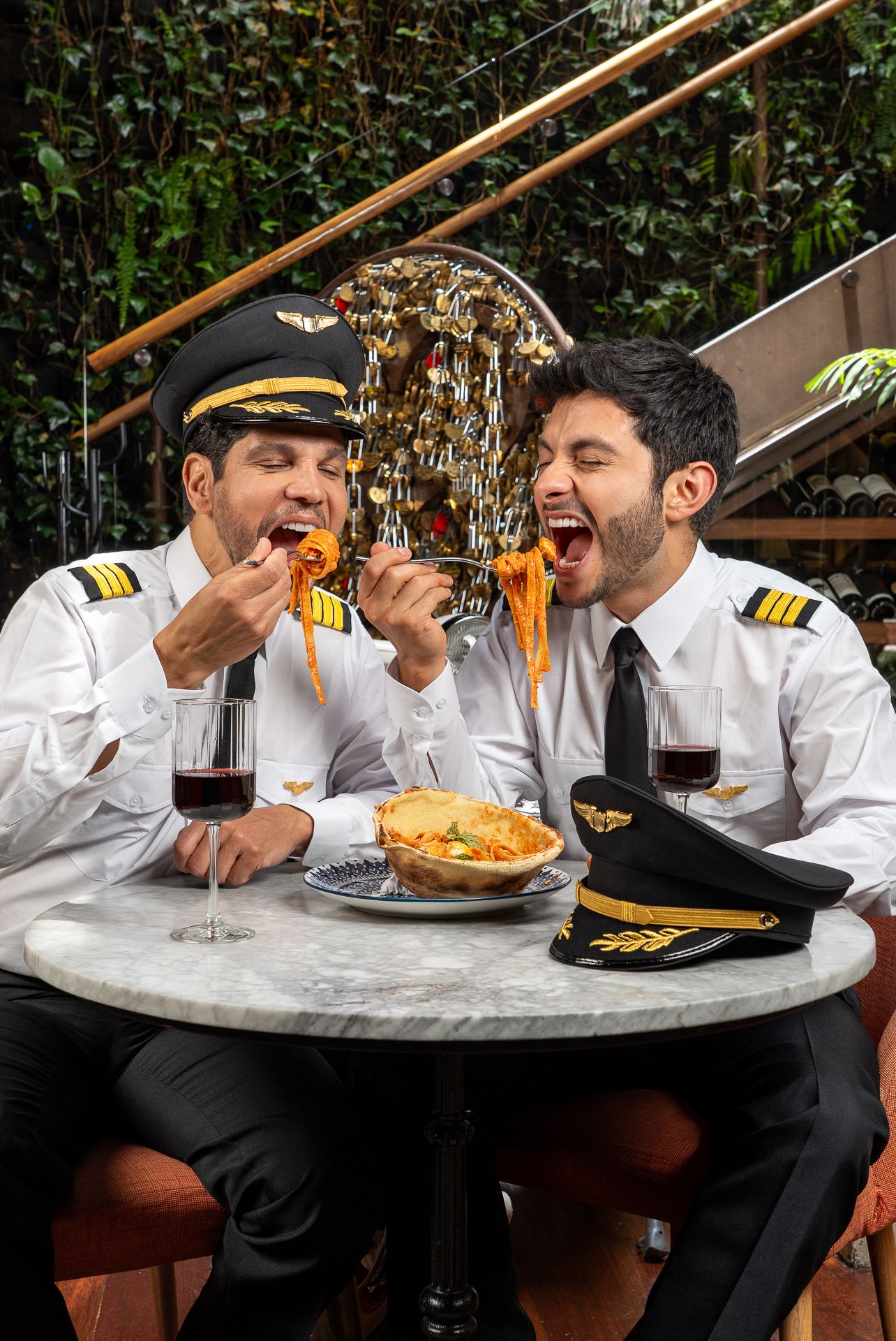 Two pilots in uniform share pasta and red wine at a marble table, laughing together in an outdoor restaurant setting.