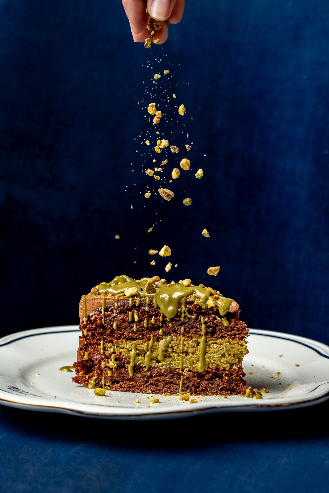 Hand sprinkling crushed pistachios over a slice of chocolate pistachio cake with green drizzle on a white plate.