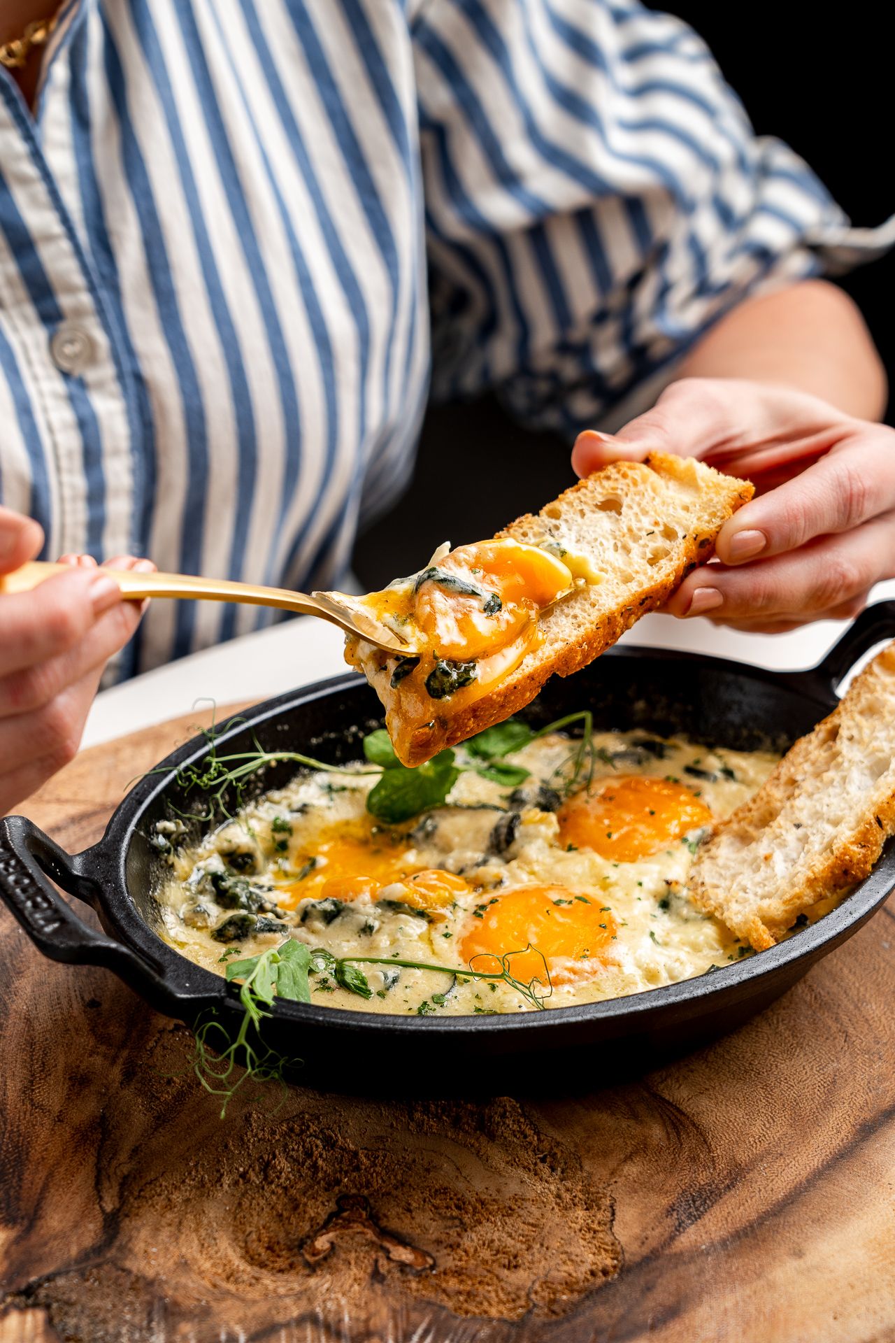 Woman dipping toast into a cast iron skillet of baked eggs with spinach and cream sauce on a wooden board.