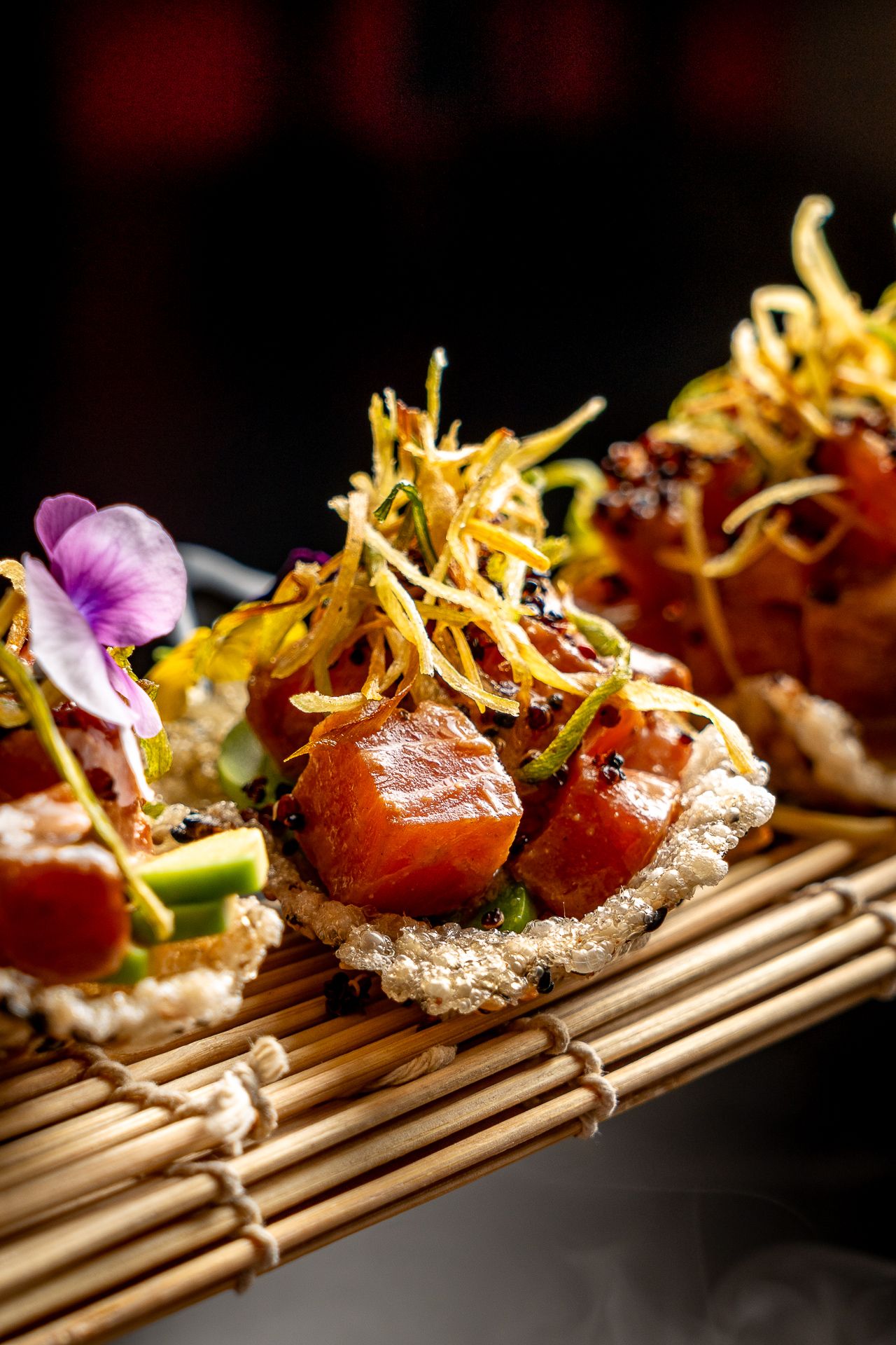 Gourmet tuna tartare bites on crispy rice topped with fried garnish and purple edible flowers on bamboo mat.