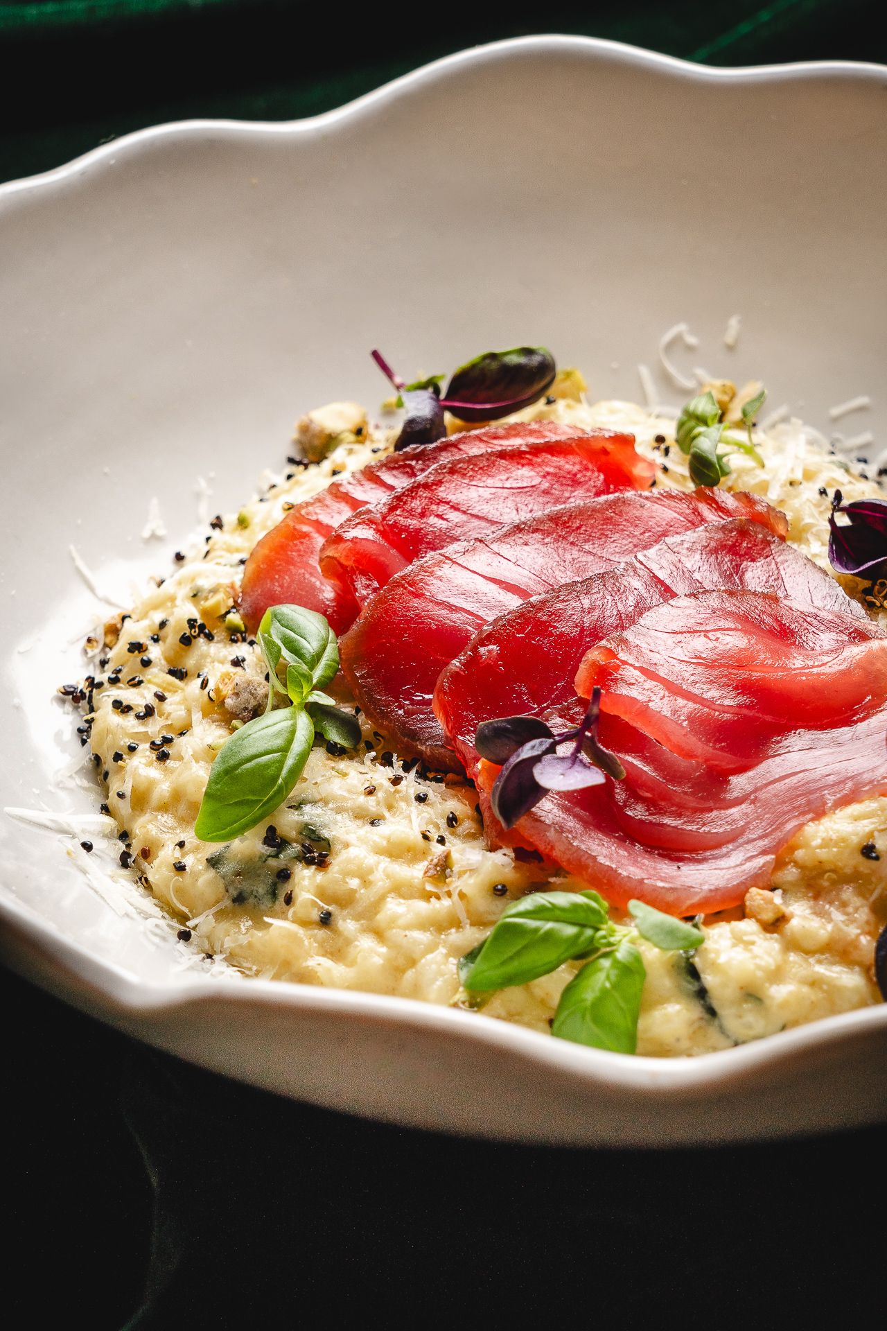 Creamy risotto topped with cured salmon, fresh basil leaves, black sesame seeds, and grated cheese in a white bowl.