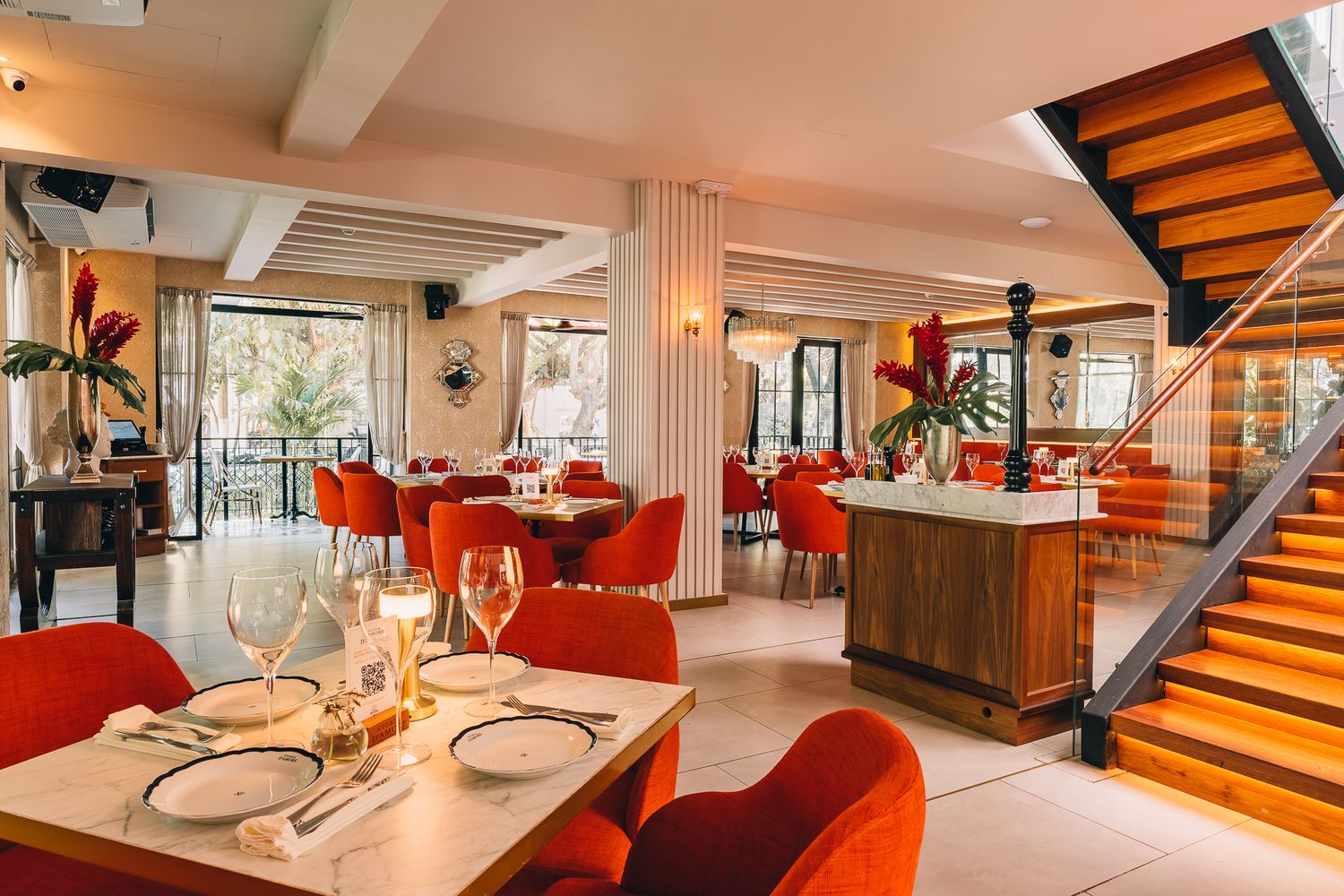 Elegant restaurant interior with red chairs, set tables, wine glasses, and a wooden staircase in a warm, modern dining space.