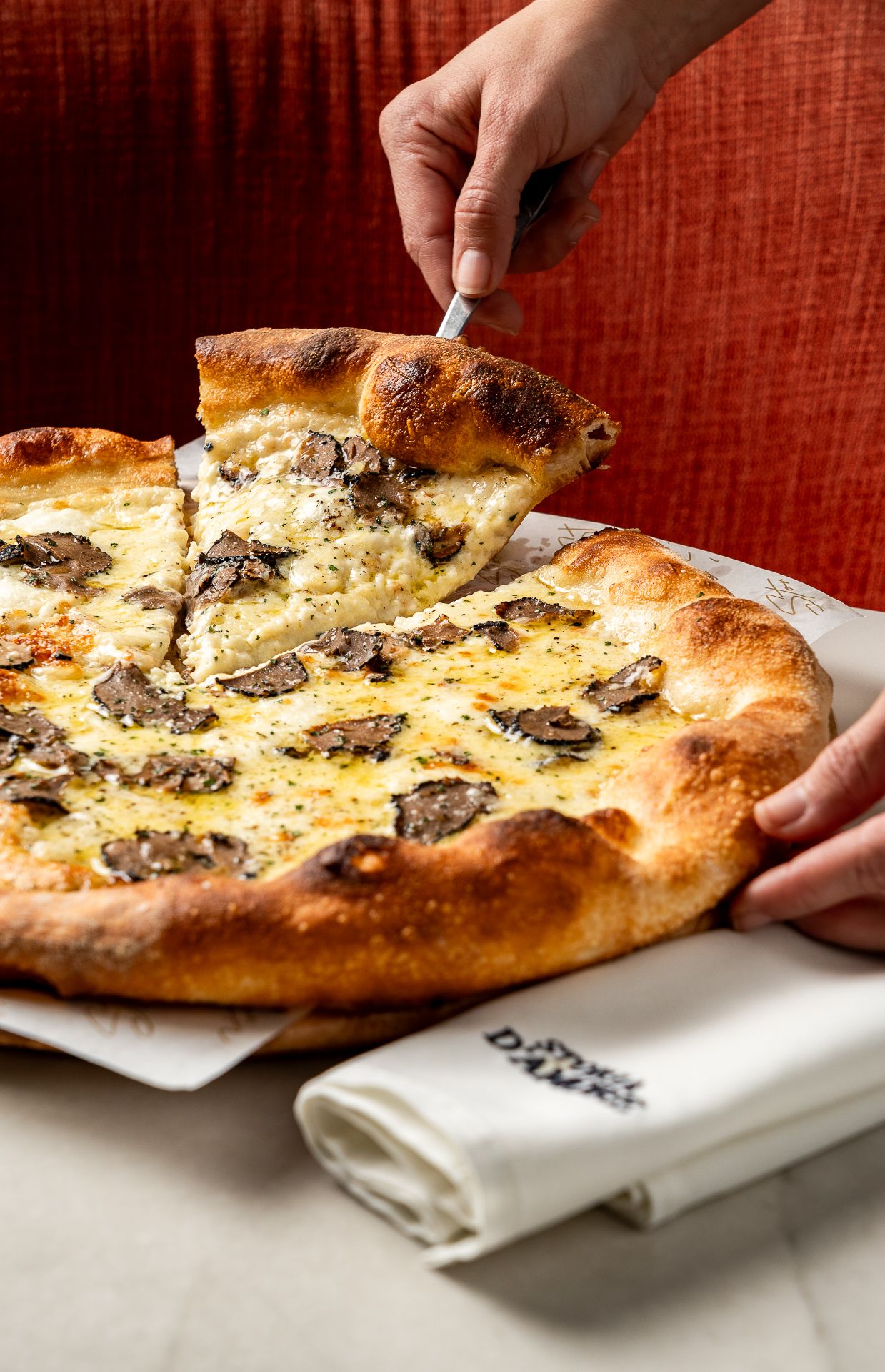 A white pizza topped with truffles and cheese being sliced, with a golden crust, served on a white napkin.