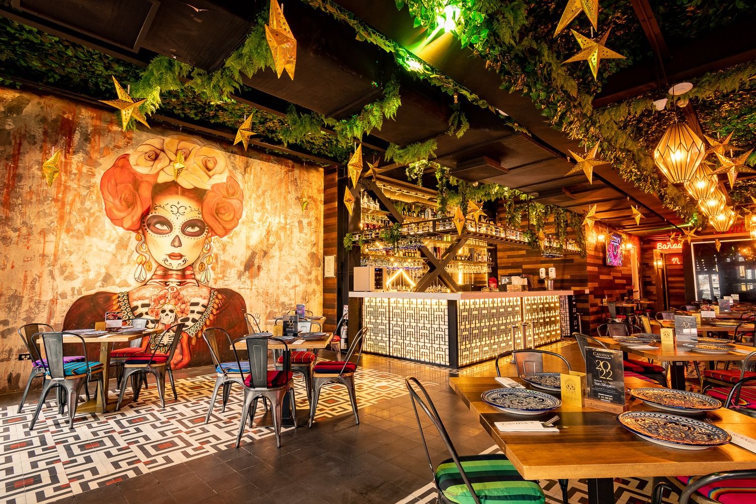 Vibrant Mexican restaurant interior with colorful murals, lush greenery, star decorations, and eclectic tiled bar area.