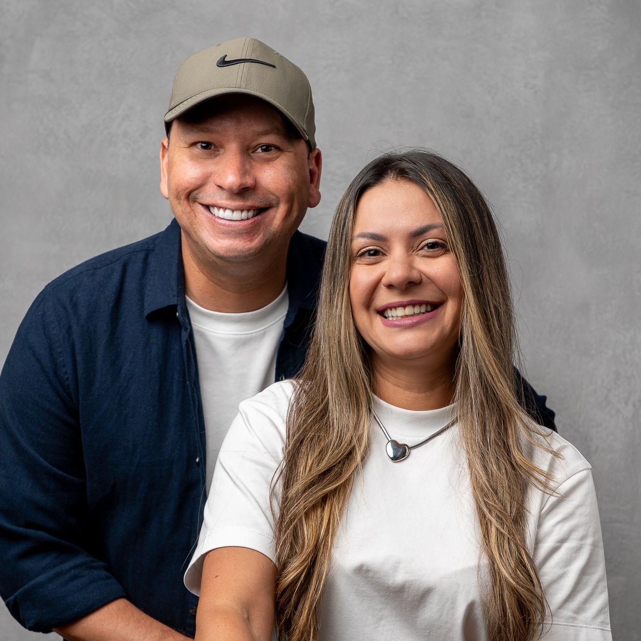A smiling man in a Nike cap and woman with long hair pose together against a gray background.