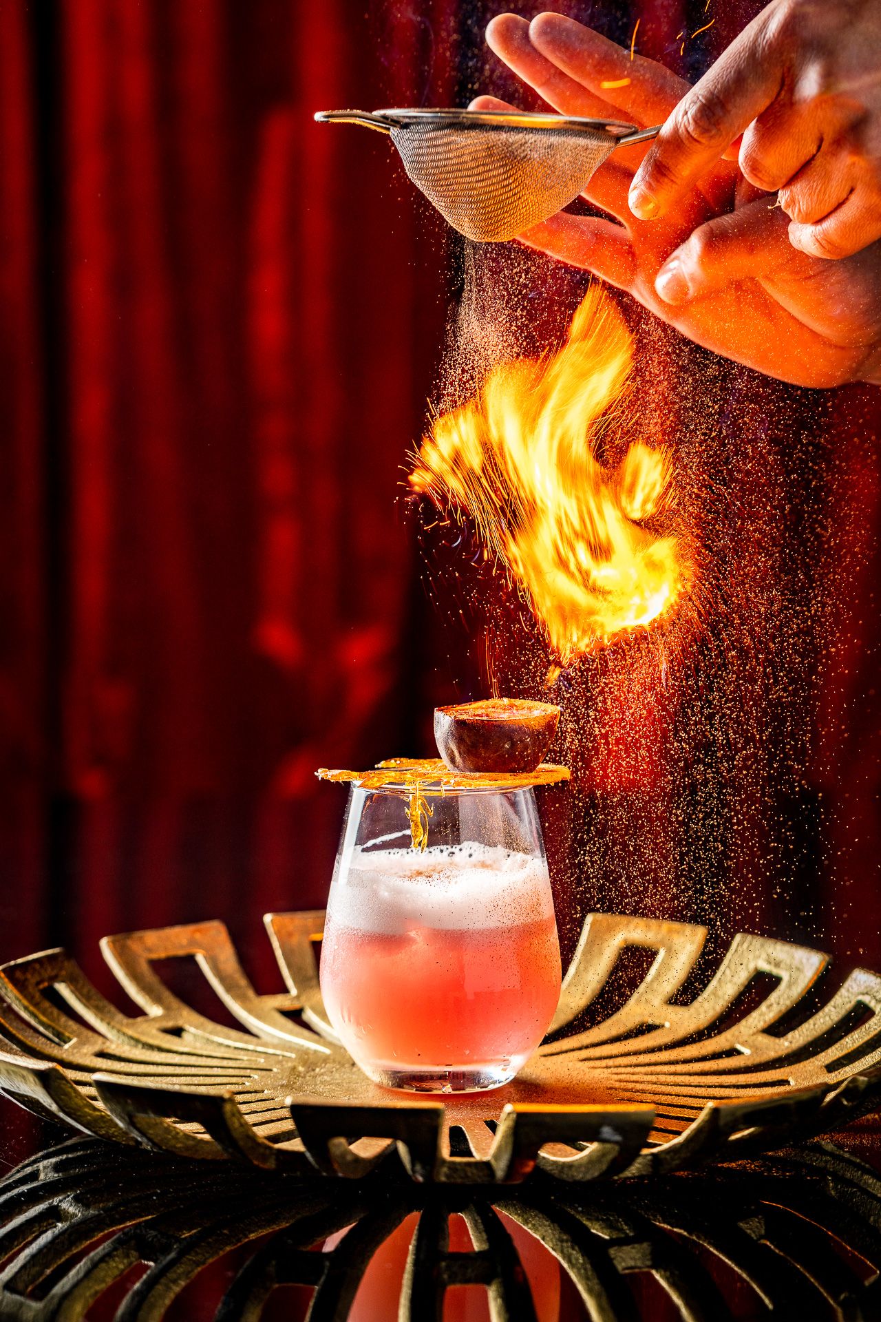 Bartender dramatically flames a cocktail garnish over a pink drink on a gold tray against a red backdrop.