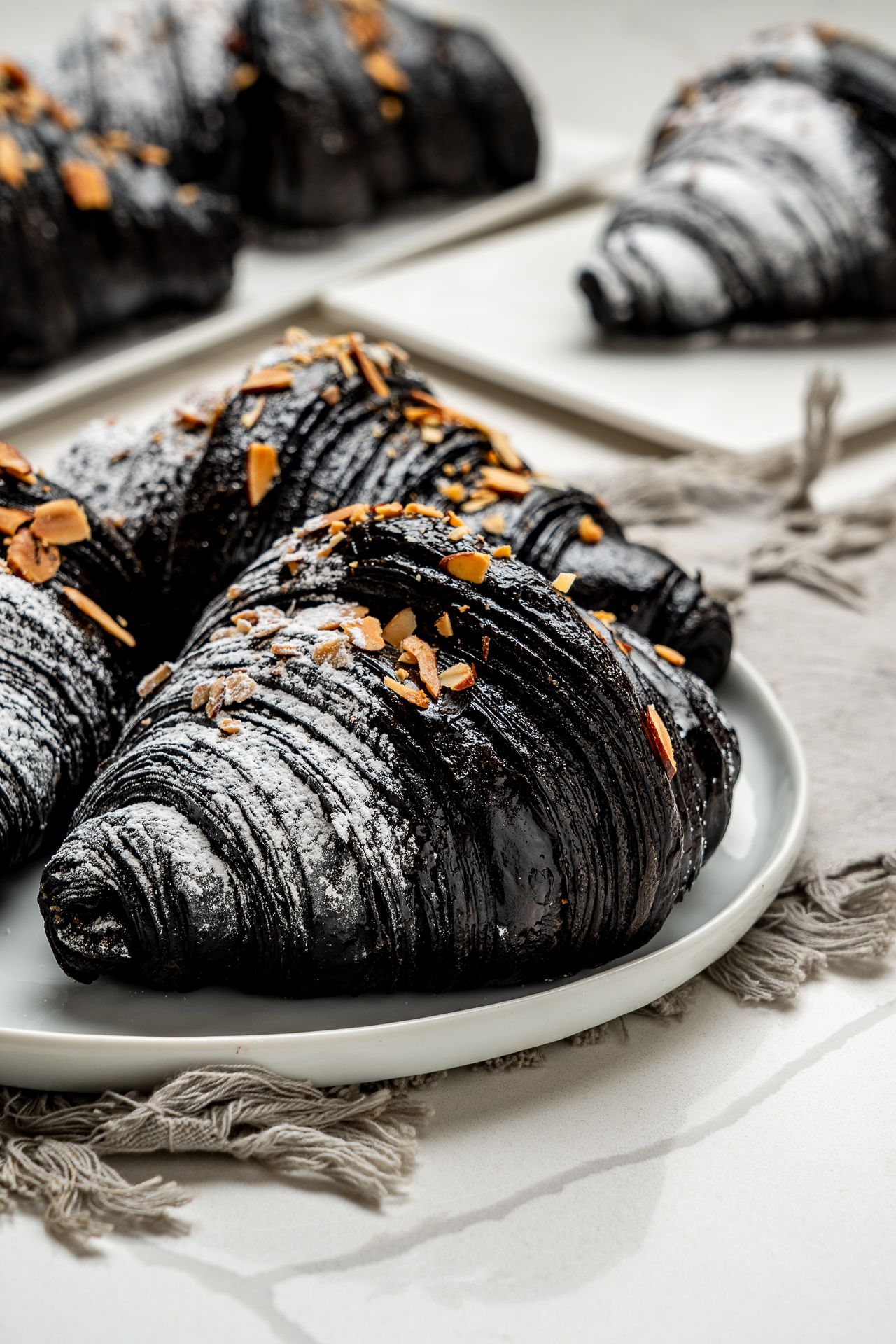 Black charcoal croissants topped with sliced almonds and powdered sugar on a white plate.