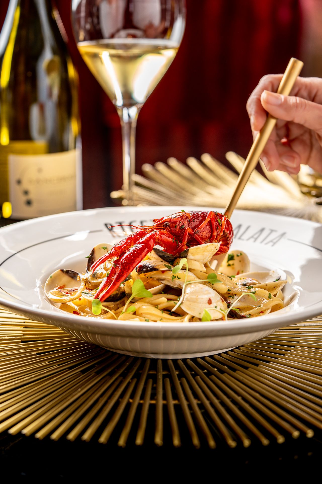 Elegant seafood pasta with red lobster and clams in a white bowl, paired with white wine in a fine dining setting.
