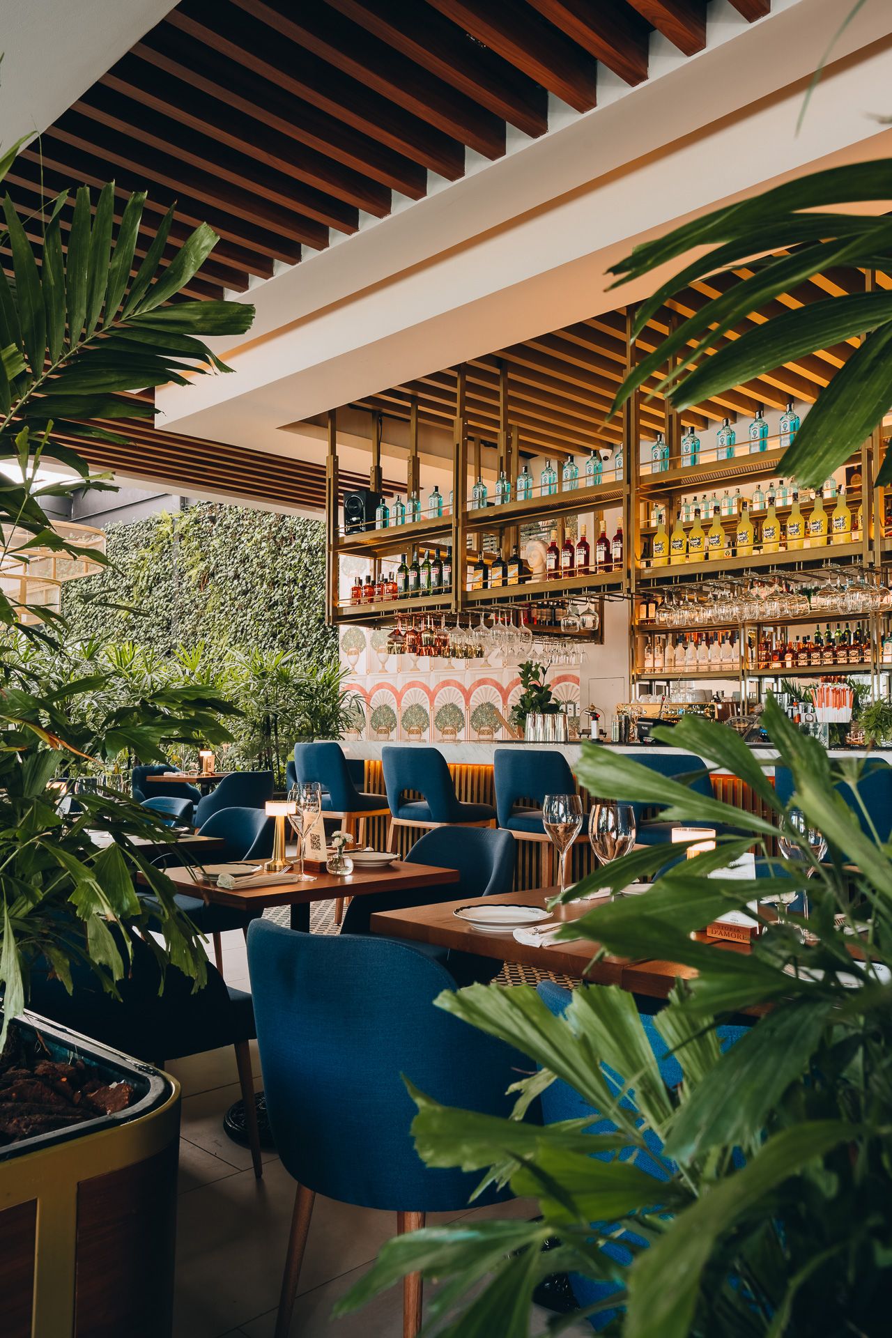 Tropical restaurant interior with blue velvet chairs, lush green plants, wooden ceiling, and a well-stocked bar with shelves of bottles.