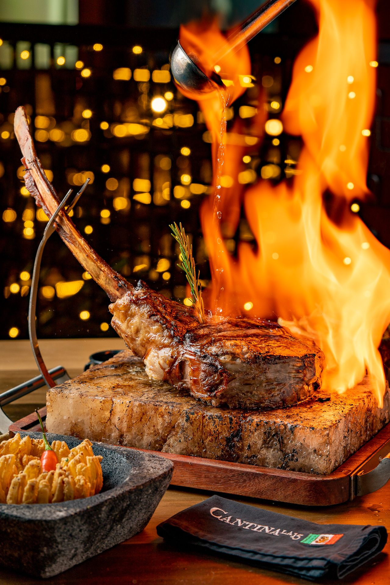 Dramatic tomahawk steak on wooden board with open flames, served at Cantina restaurant with sides and bokeh lights.