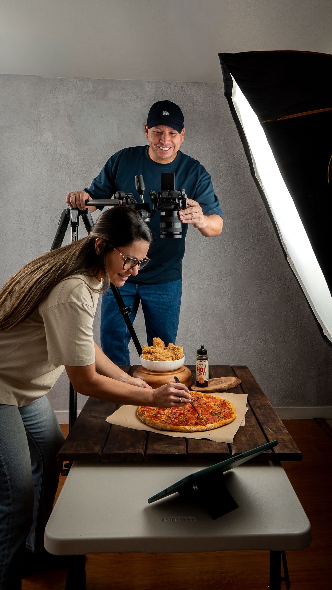 Food photographers styling a pizza and fried chicken in a professional studio with softbox lighting and camera equipment.