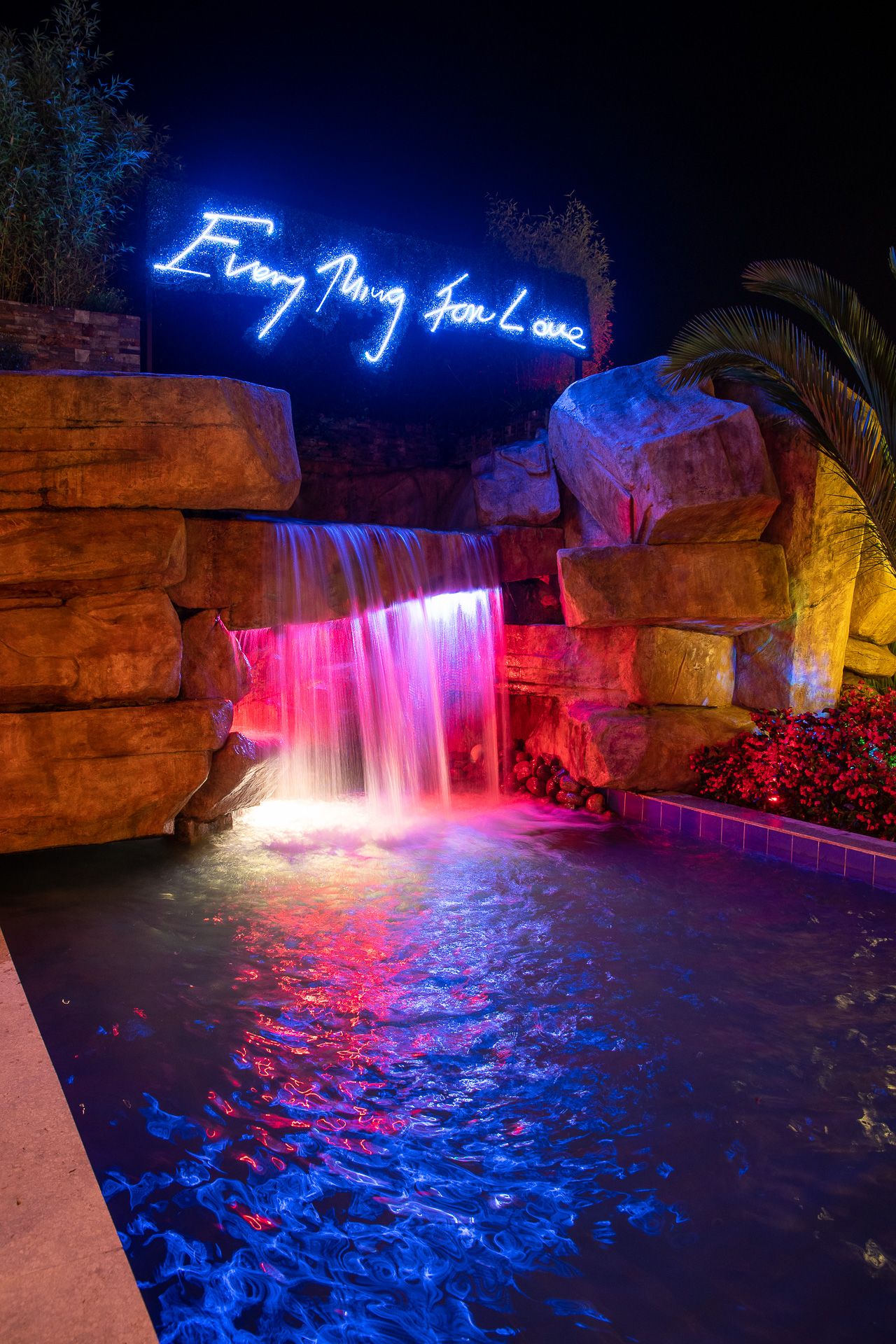 Neon sign reading Everything for Love above a colorful waterfall with pink and blue lights reflecting on a pool at night.