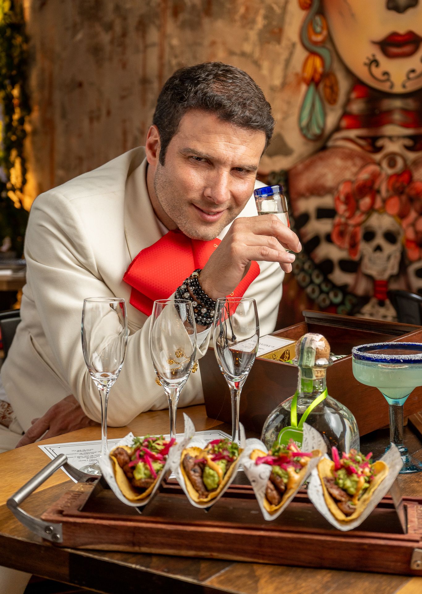 Man in white suit with red tie examines tequila shot beside tacos and cocktails at a Mexican restaurant.