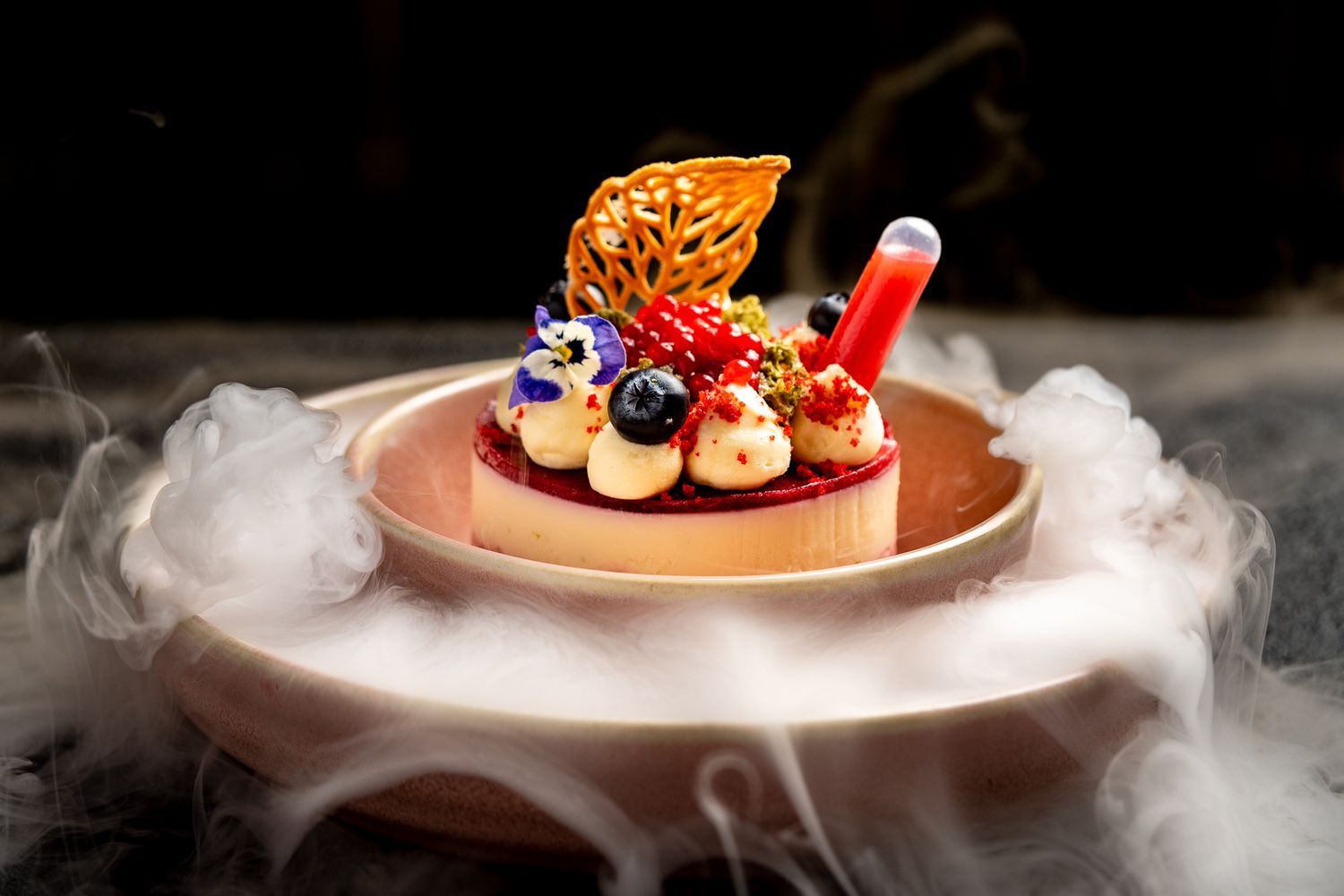 Elegant dessert tart topped with berries, cream, edible gold, and flowers, surrounded by dramatic dry ice smoke on a wooden plate.