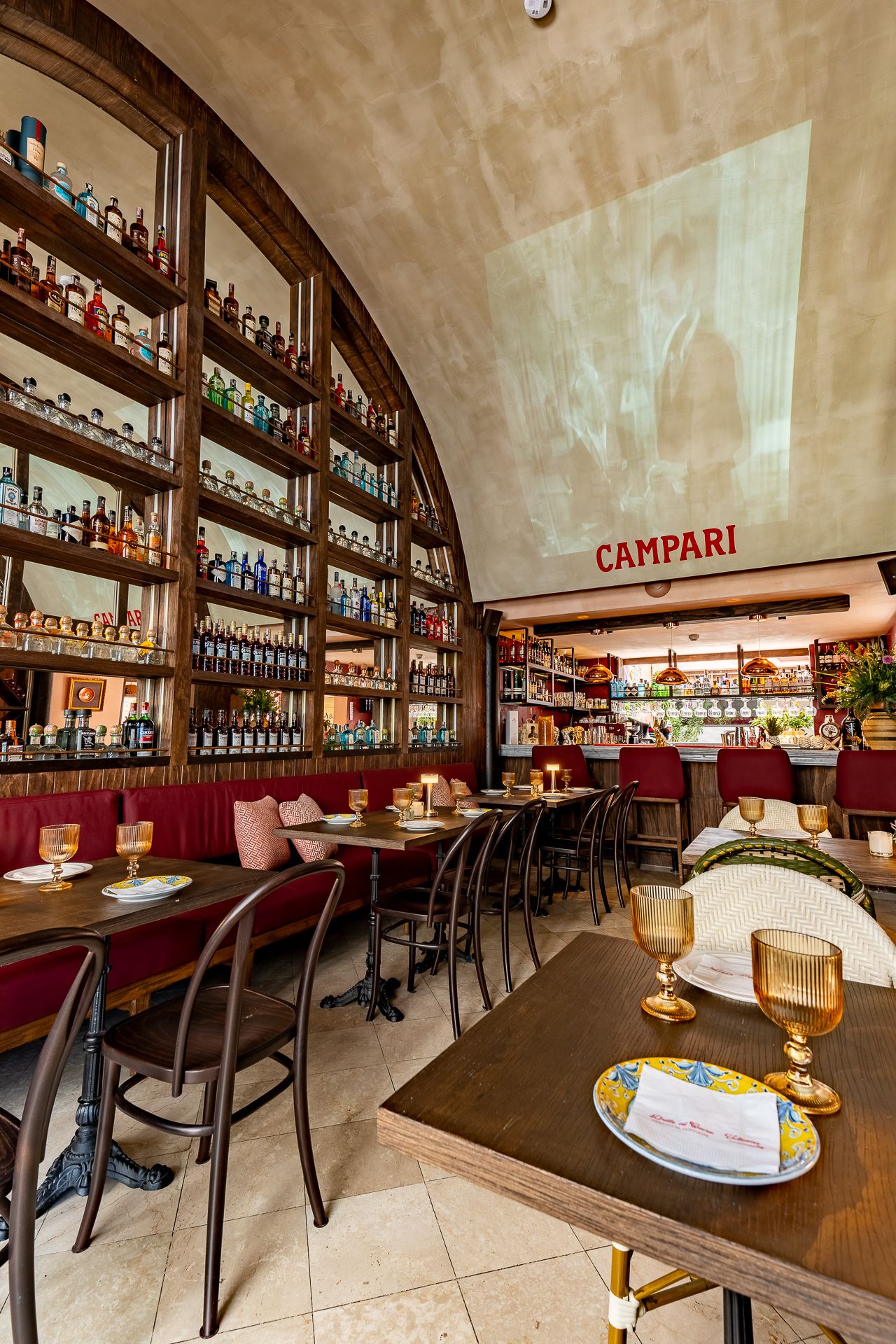 Elegant Italian restaurant interior with arched shelves of liquor bottles, red banquettes, and a glowing Campari sign above the bar.