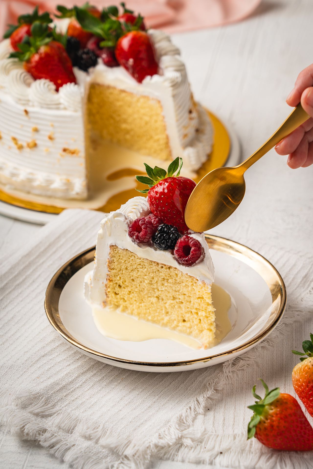 A slice of vanilla layer cake with white frosting and fresh berries served on a plate, with the full cake in the background.
