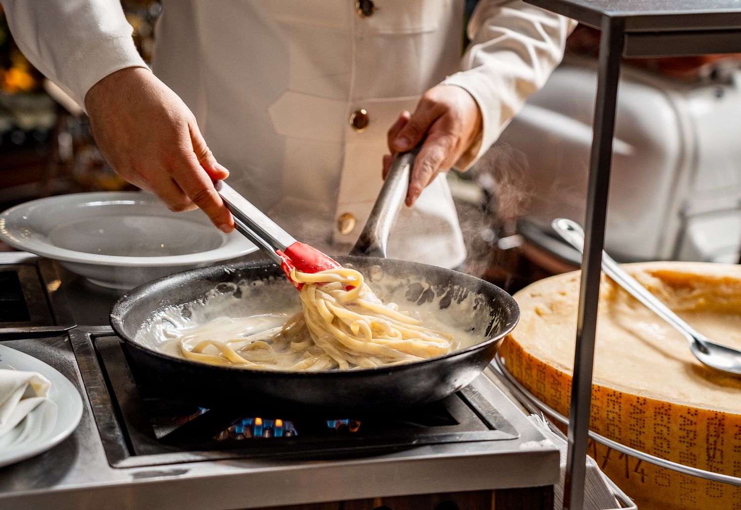 Chef tossing fresh pasta in a pan on a gas stove using red-tipped tongs in a professional kitchen.