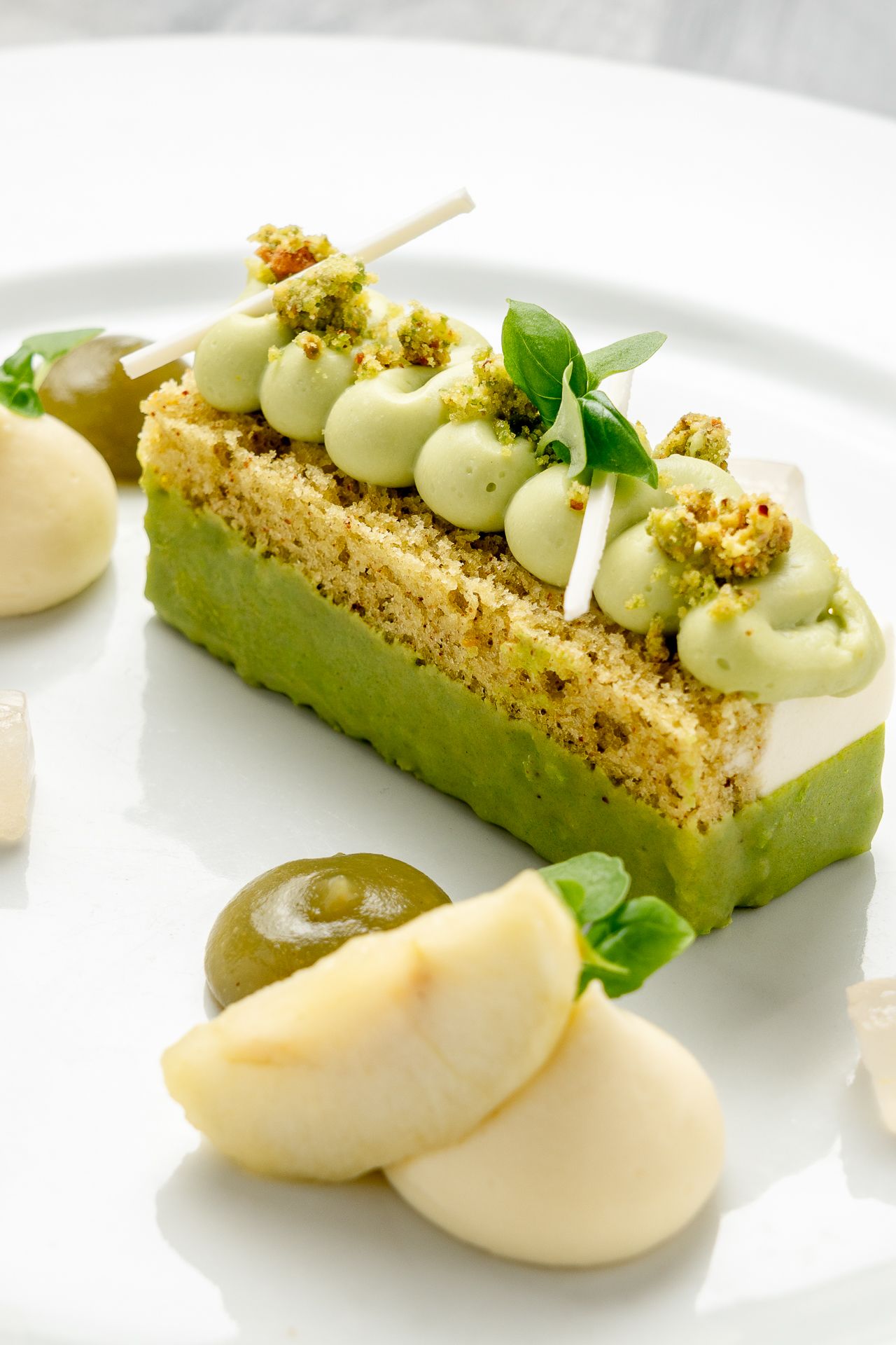 Elegant pistachio dessert bar with green mousse, crumble topping, fresh basil, and poached pear slices on white plate.