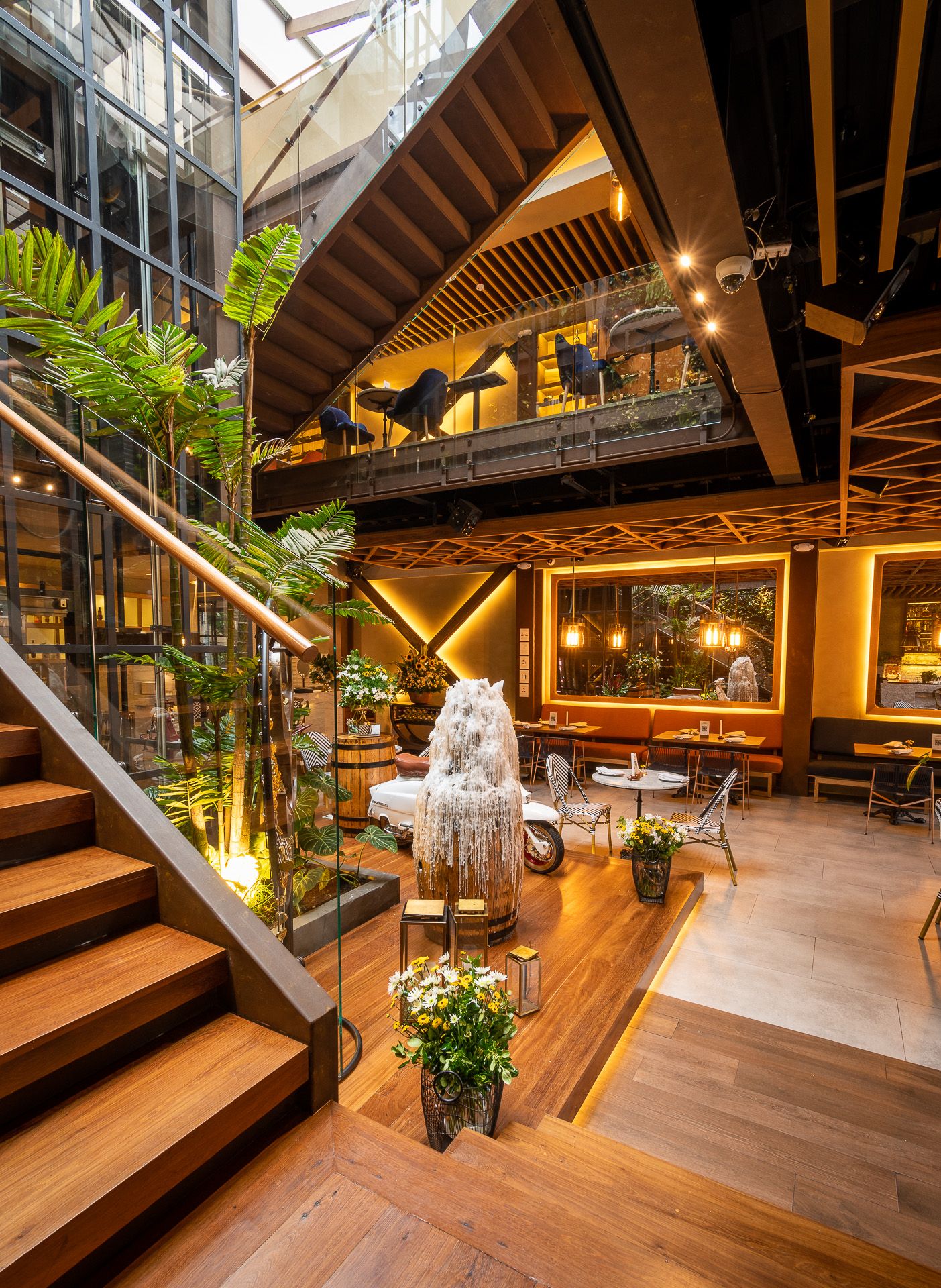 Modern multi-level restaurant interior with wooden stairs, lush tropical plants, warm lighting, and elegant dining area.