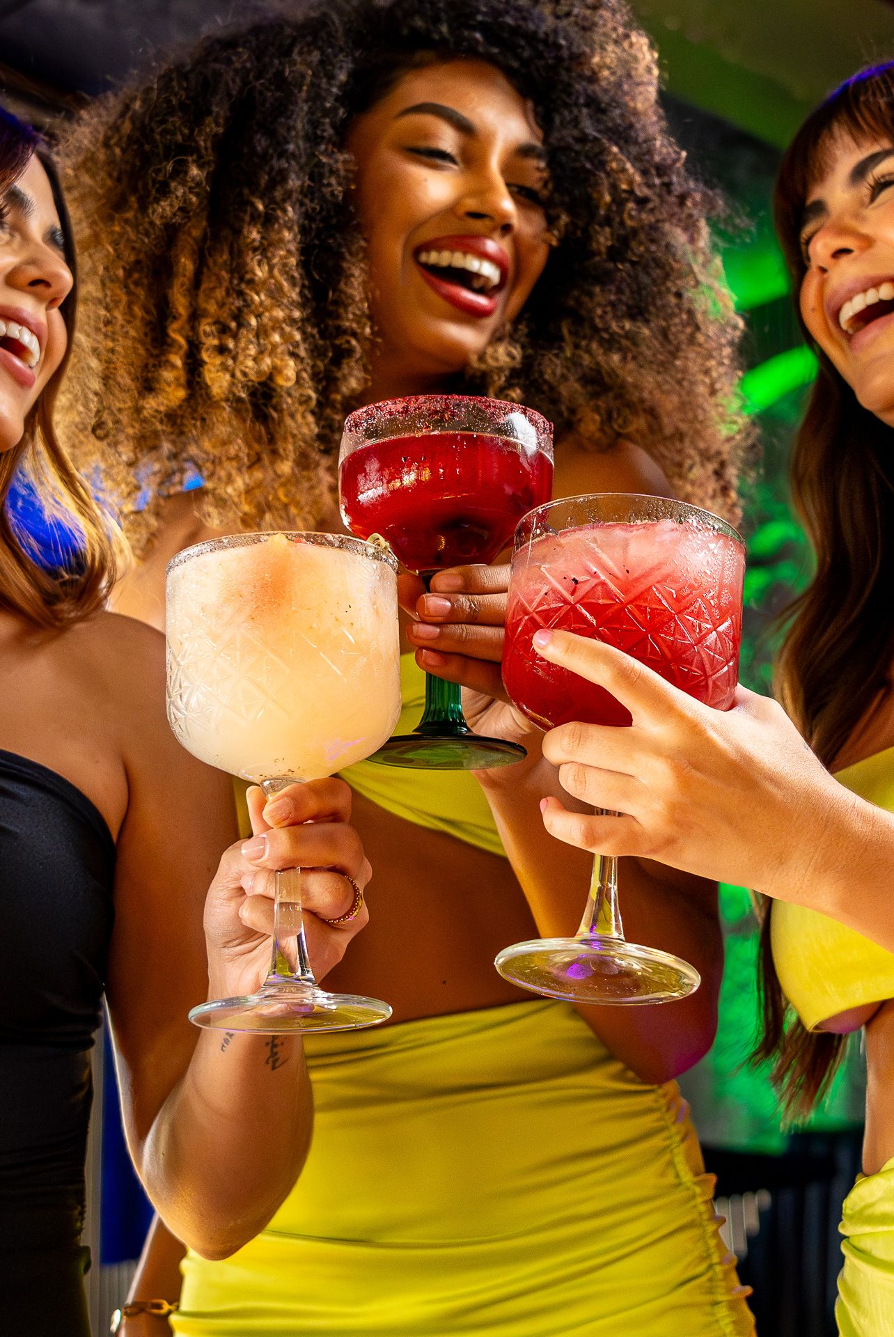 Three women laughing and toasting with colorful cocktails at a vibrant nightclub party.