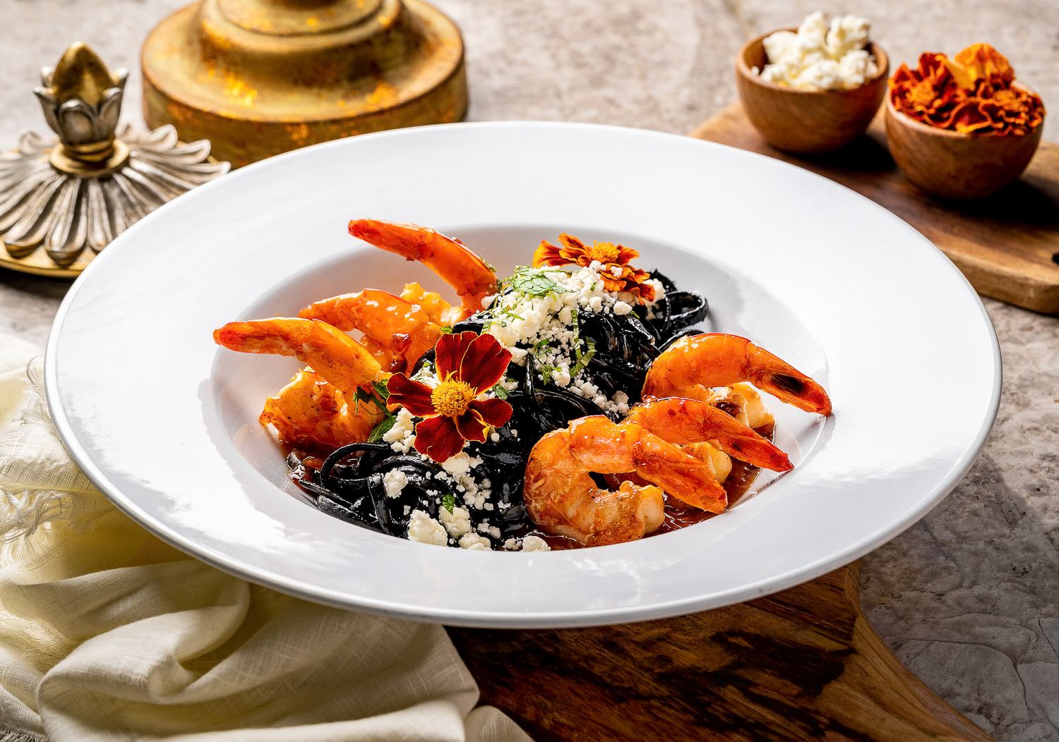 Black squid ink pasta with large shrimp, crumbled cheese, and crispy bacon in a white bowl.