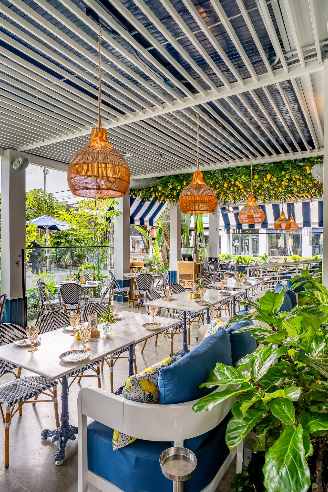 Bright restaurant patio with wicker pendant lights, blue cushioned seating, marble tables, striped awnings, and lush tropical plants.