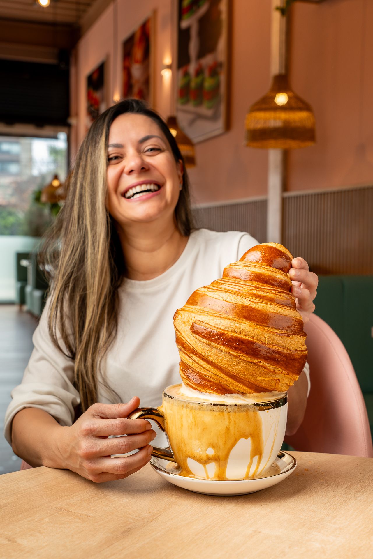 Smiling woman holding a giant croissant on a coffee cup at a cozy cafe.