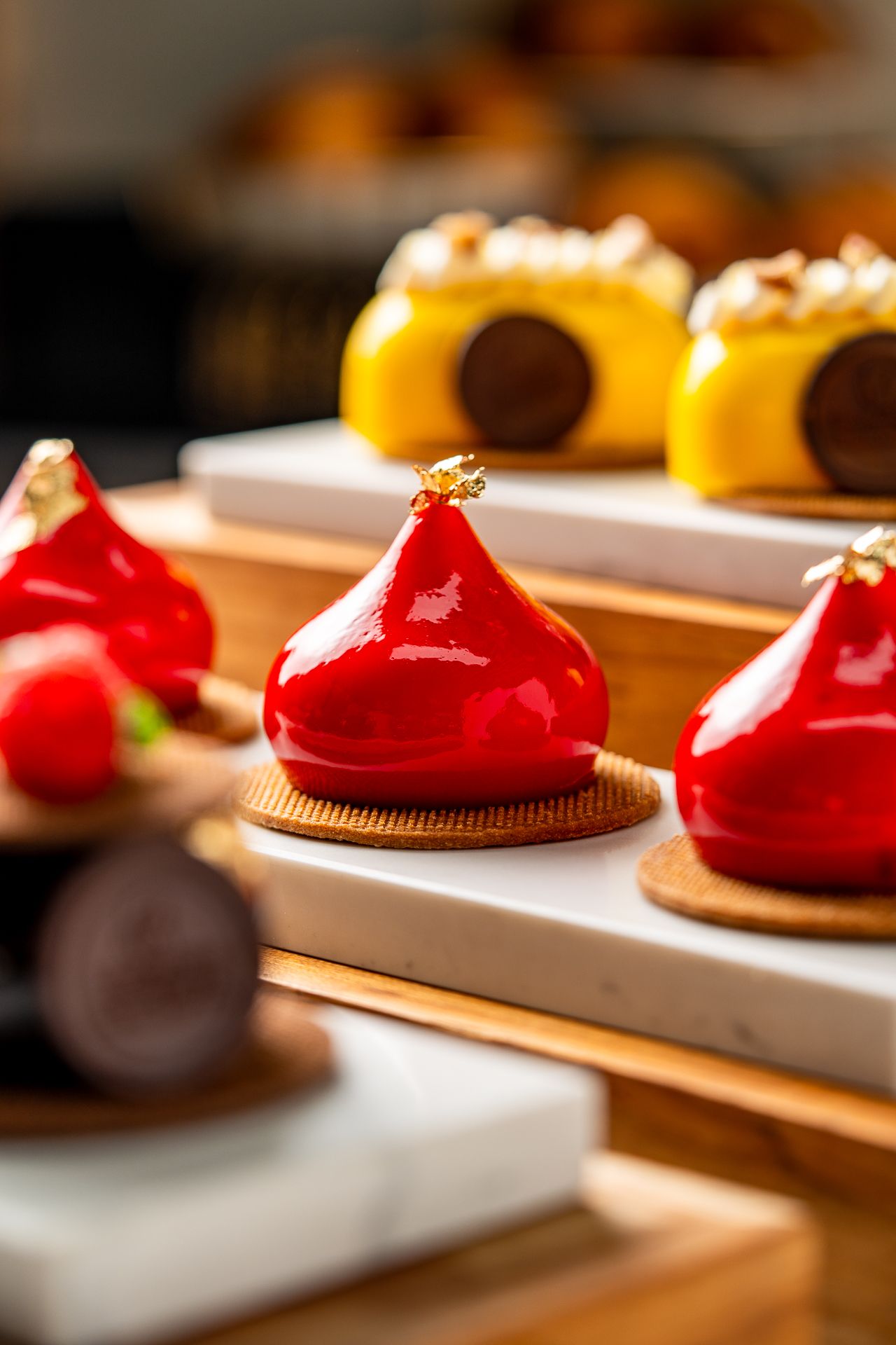 Elegant glossy red and yellow mirror-glazed petit gateaux desserts displayed on marble and wood stands.