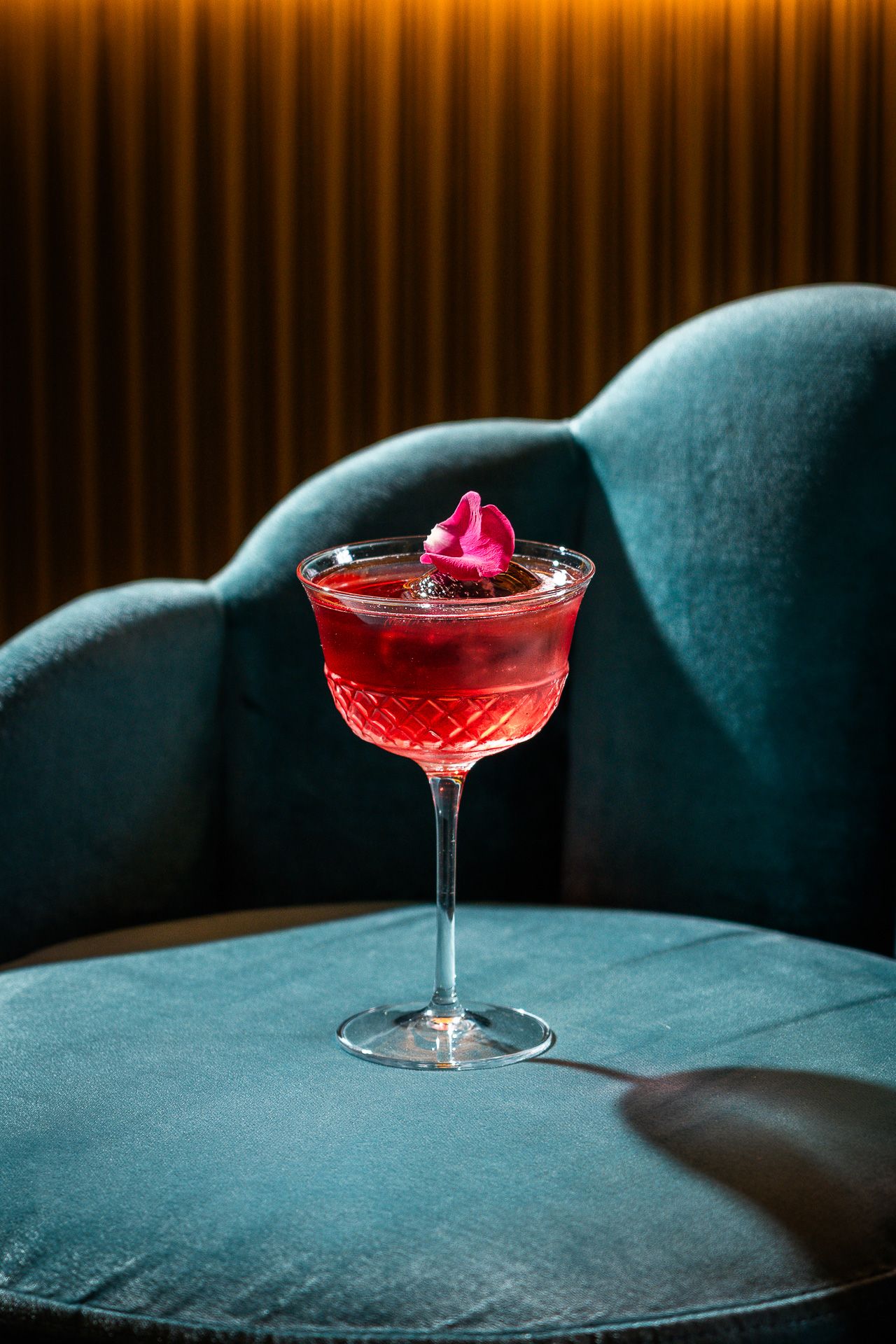A vibrant red cocktail in a crystal coupe glass, garnished with a pink flower, resting on a teal velvet chair.