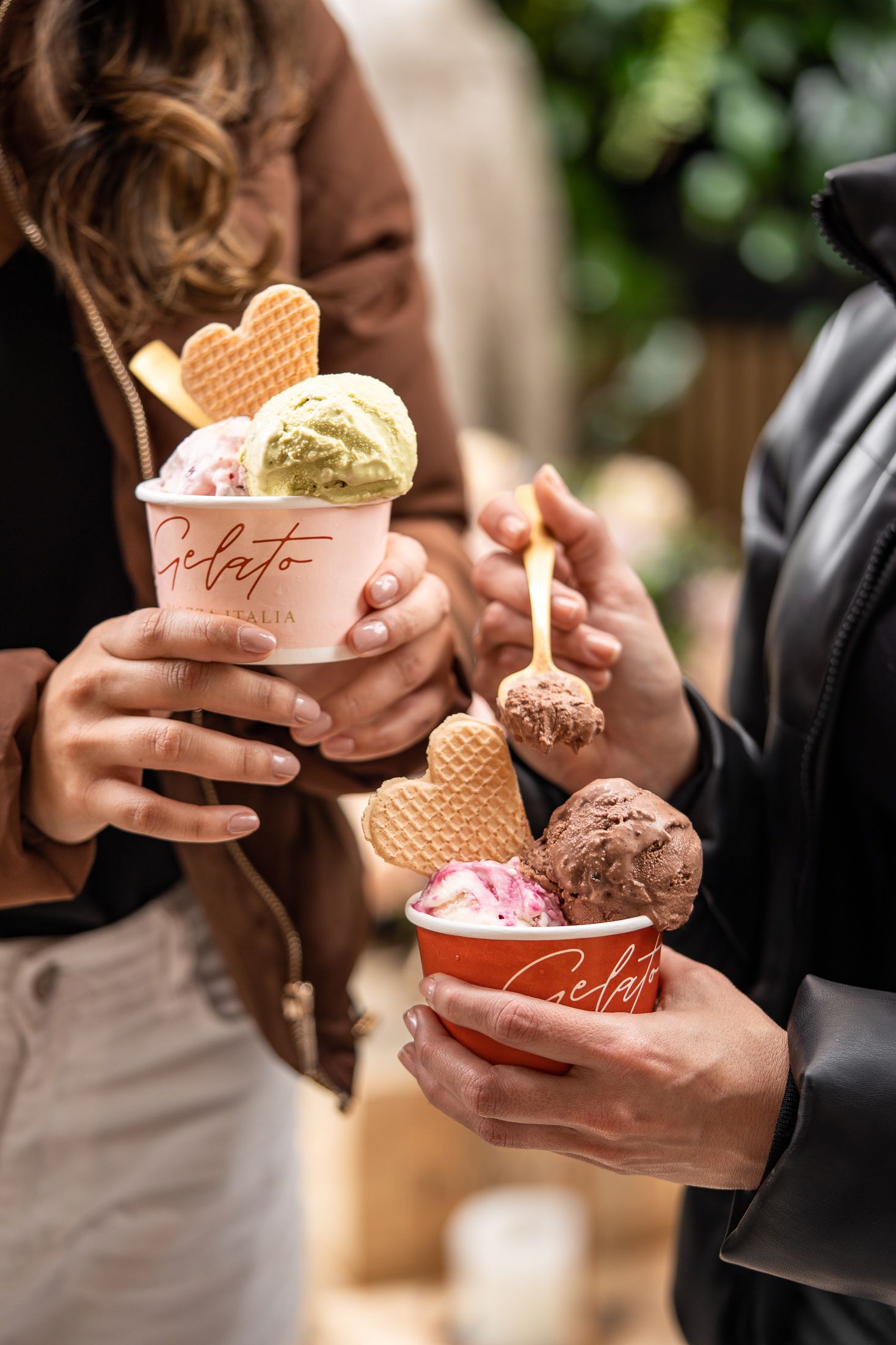 Two people holding Gelato Italia cups filled with colorful ice cream scoops and heart-shaped waffle cookies outdoors.