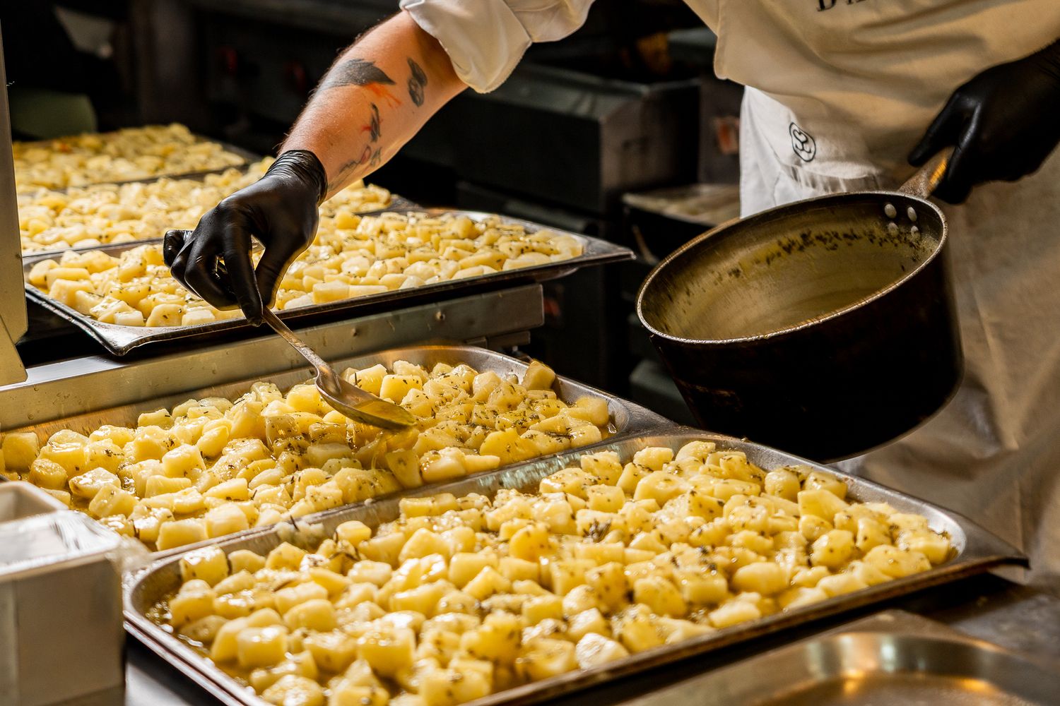 Chef preparing gnocchi on large baking trays in a professional kitchen, wearing black gloves and white uniform.