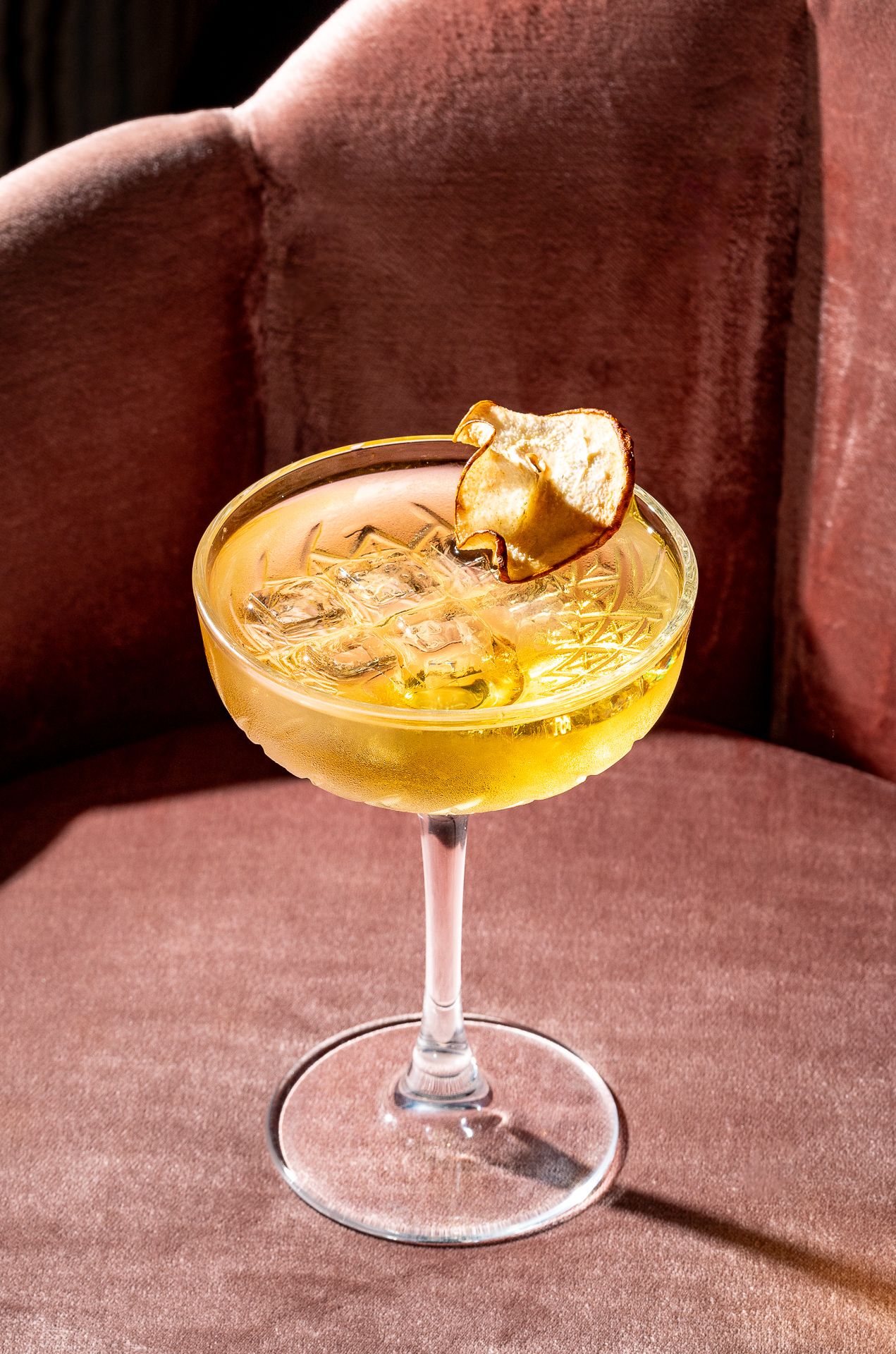 Elegant golden cocktail in a coupe glass garnished with a dried fruit slice, served on a pink velvet chair.