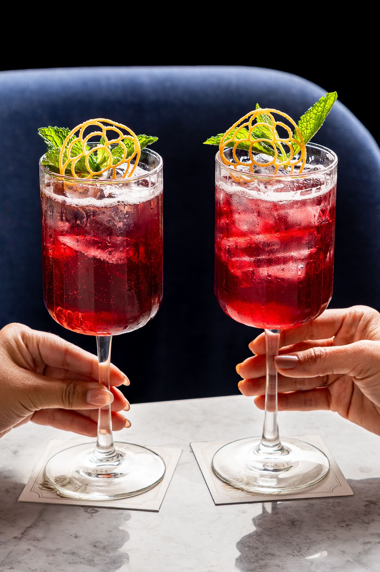 Two vibrant red cocktails garnished with mint and lemon zest on a marble table.