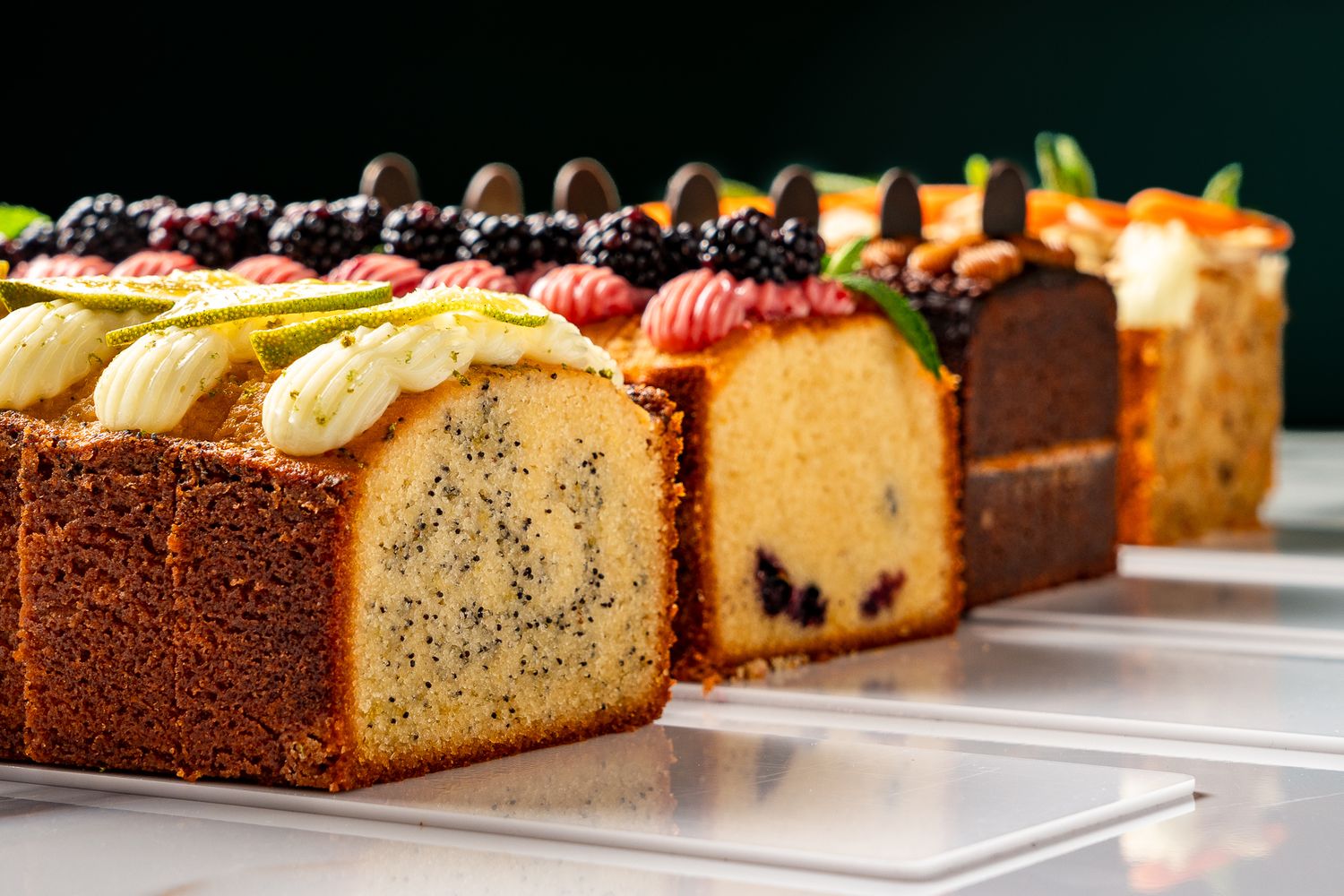 Assorted gourmet loaf cakes topped with fresh berries and cream displayed in a elegant row on a white surface.