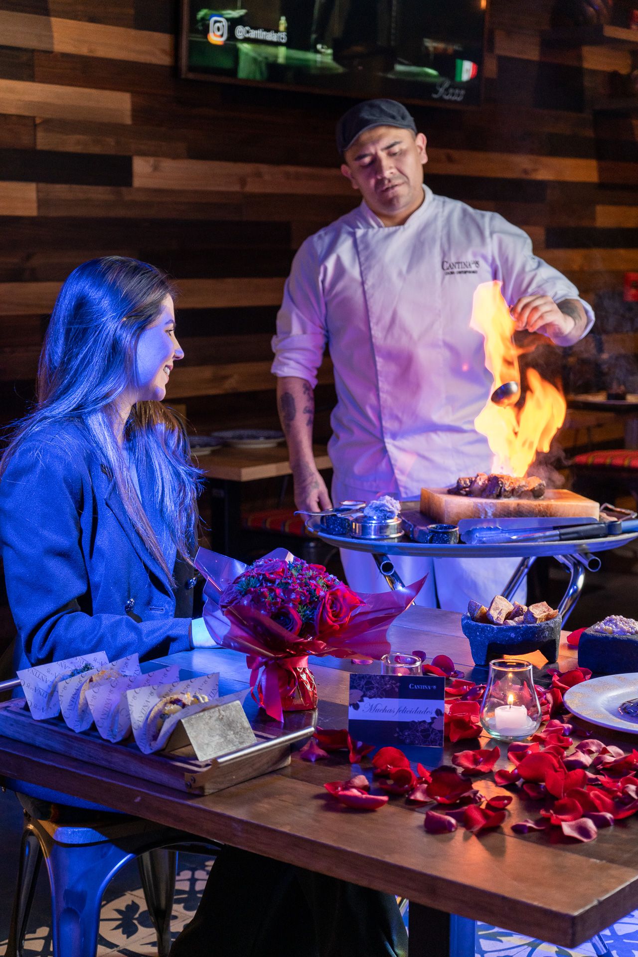 Chef flambeing a dish tableside for a woman at a romantic restaurant decorated with rose petals and flowers.