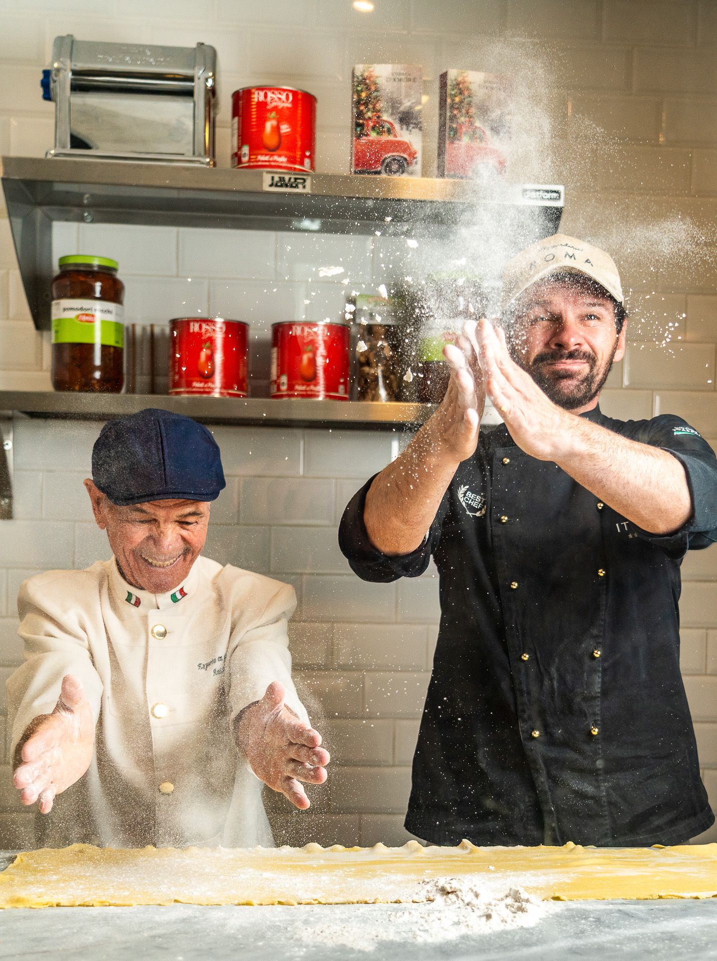 Two chefs joyfully tossing flour over fresh pasta dough in a professional kitchen with spices on shelves.